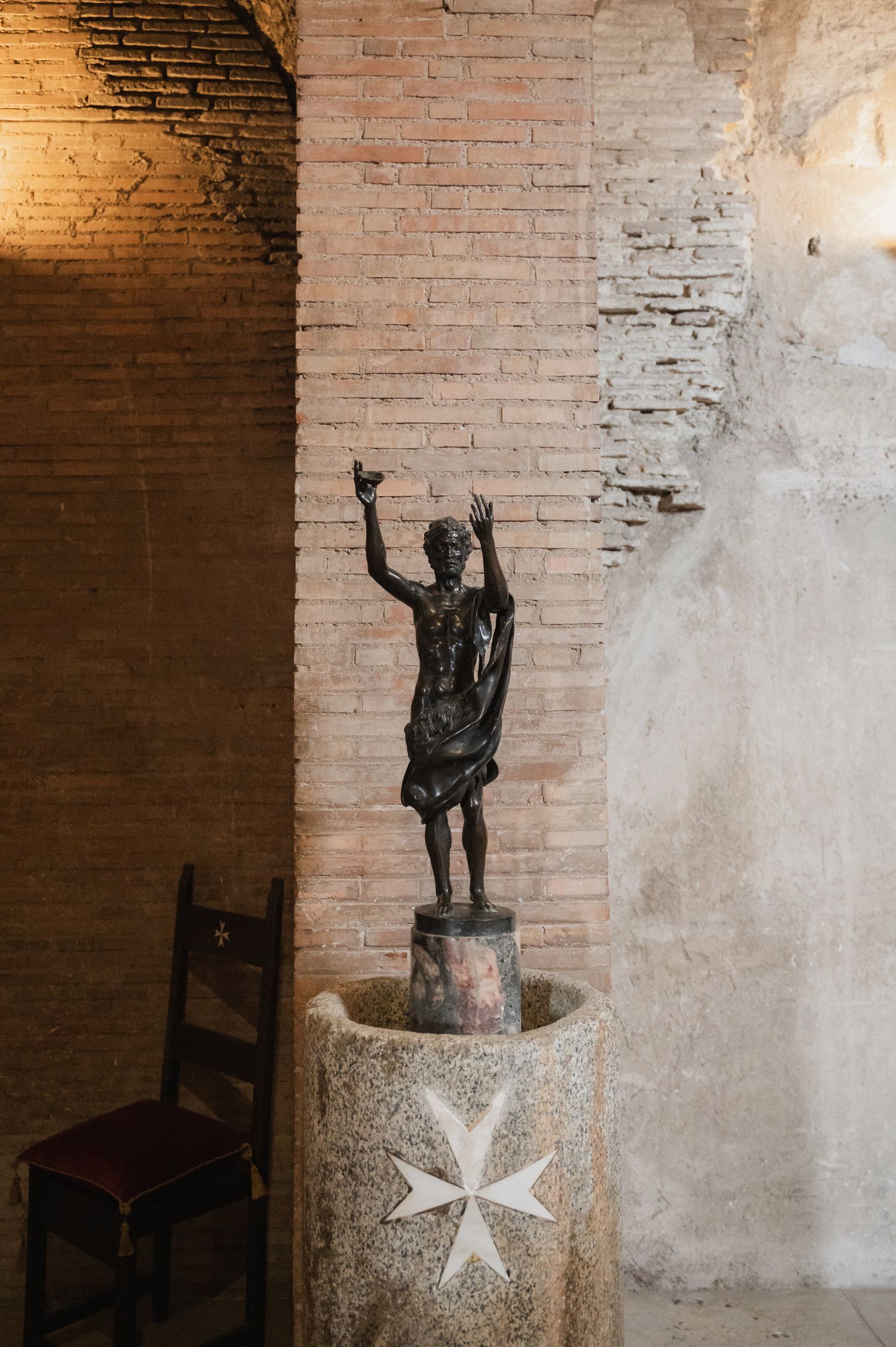 Small bronze statue on a stone pedestal with a Maltese cross symbol against a rustic brick wall.