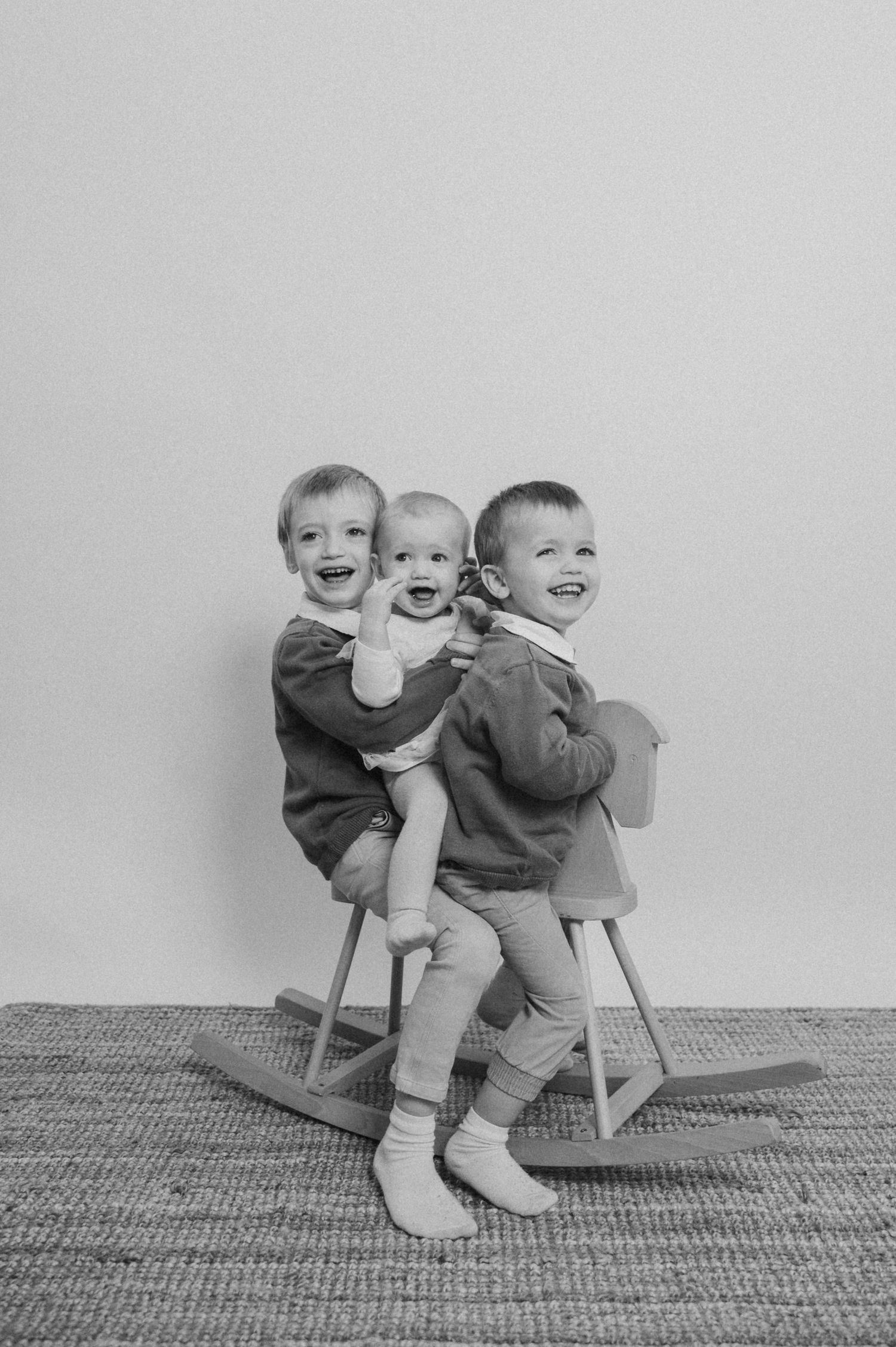 Two children sit playfully on wooden chairs against a plain wall in a black and white photograph.