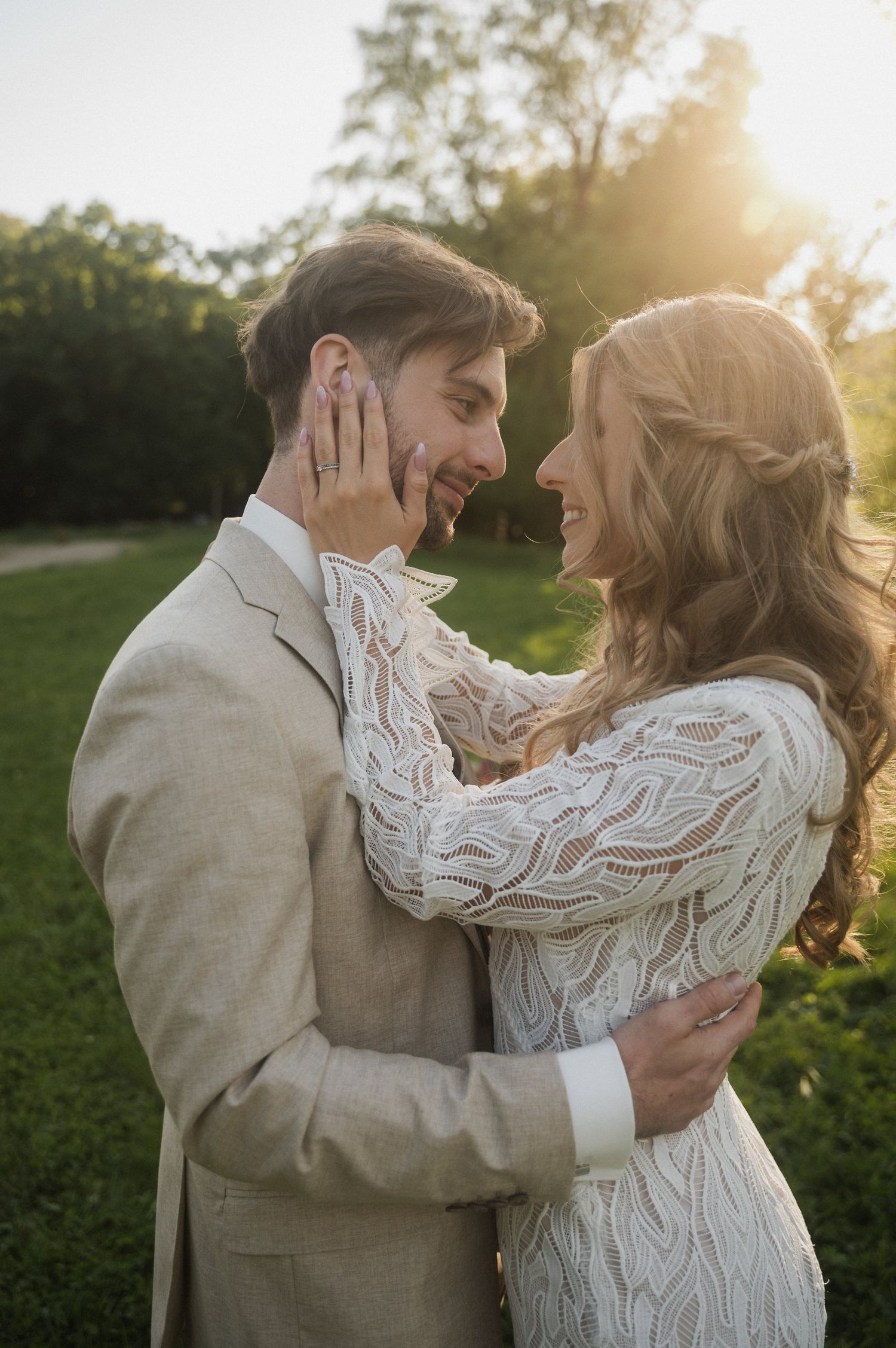 A romantic couple embraces in a sunlit meadow at golden hour, with the bride wearing a lace long-sleeved dress.