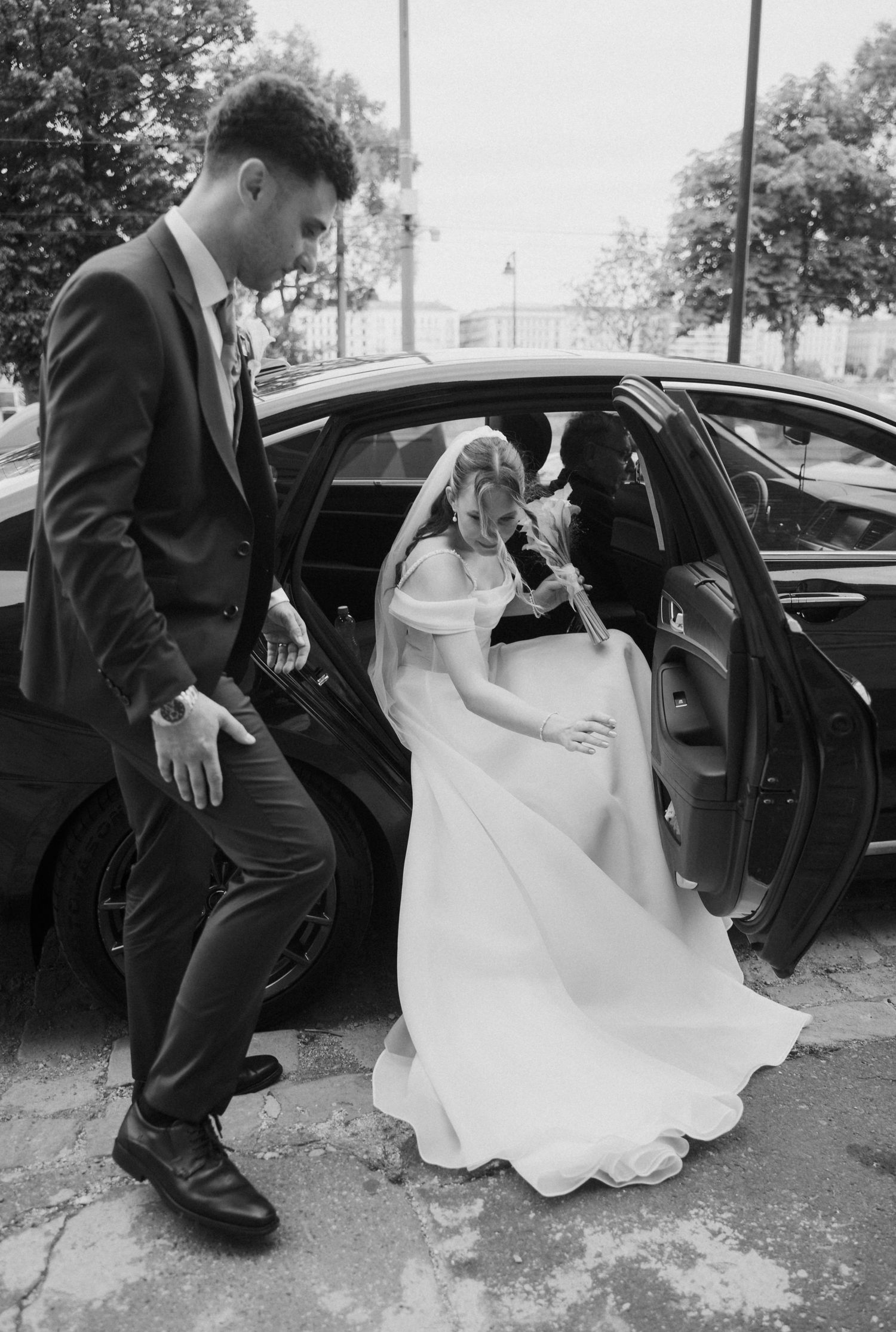 Black and white photo of groom helping bride exit a luxury car in her flowing wedding dress.