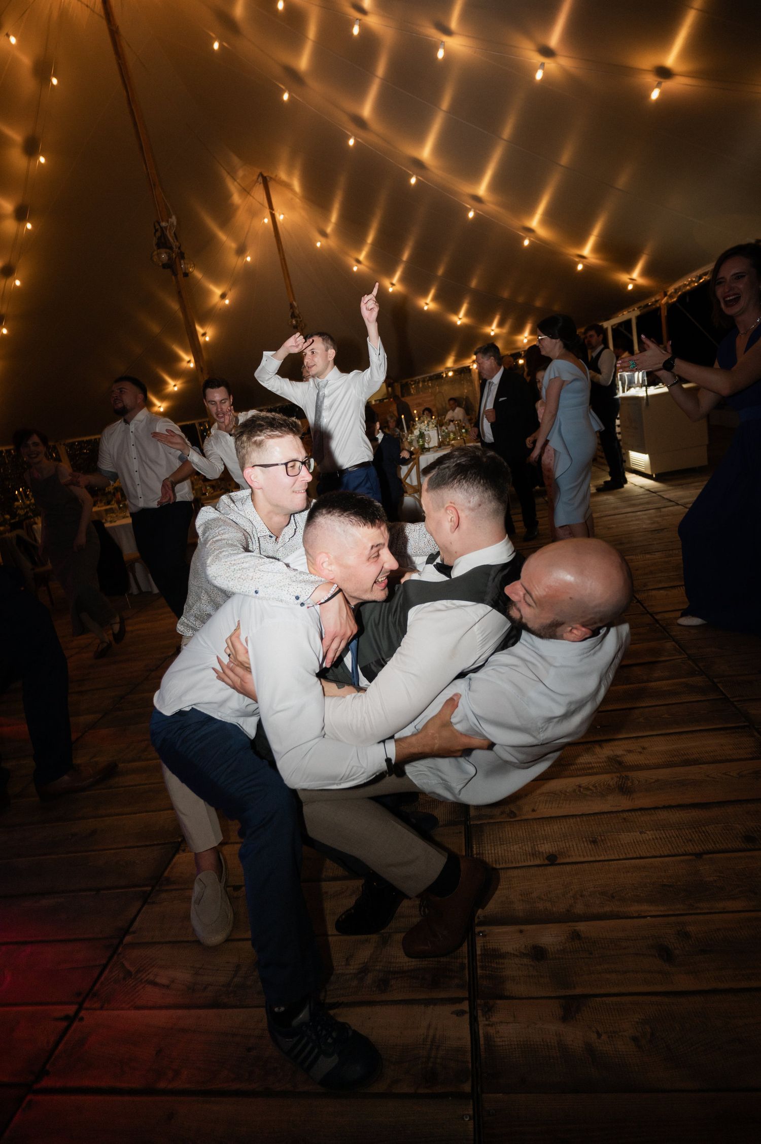 Two people dance energetically at a wedding reception under warm lighting in a tent venue.