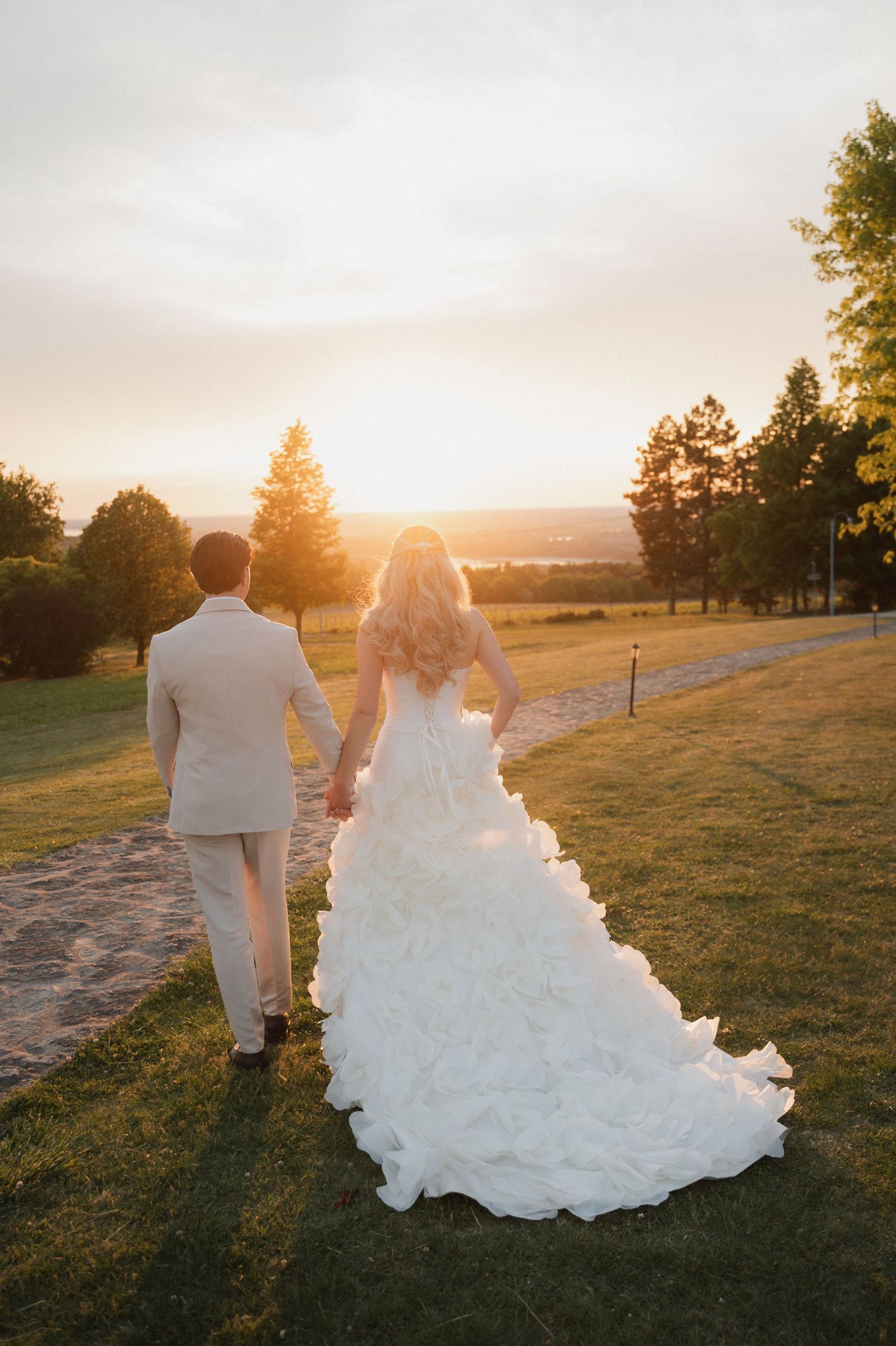 Bride and groom walking hand in hand toward golden sunset on lush green wedding venue grounds.
