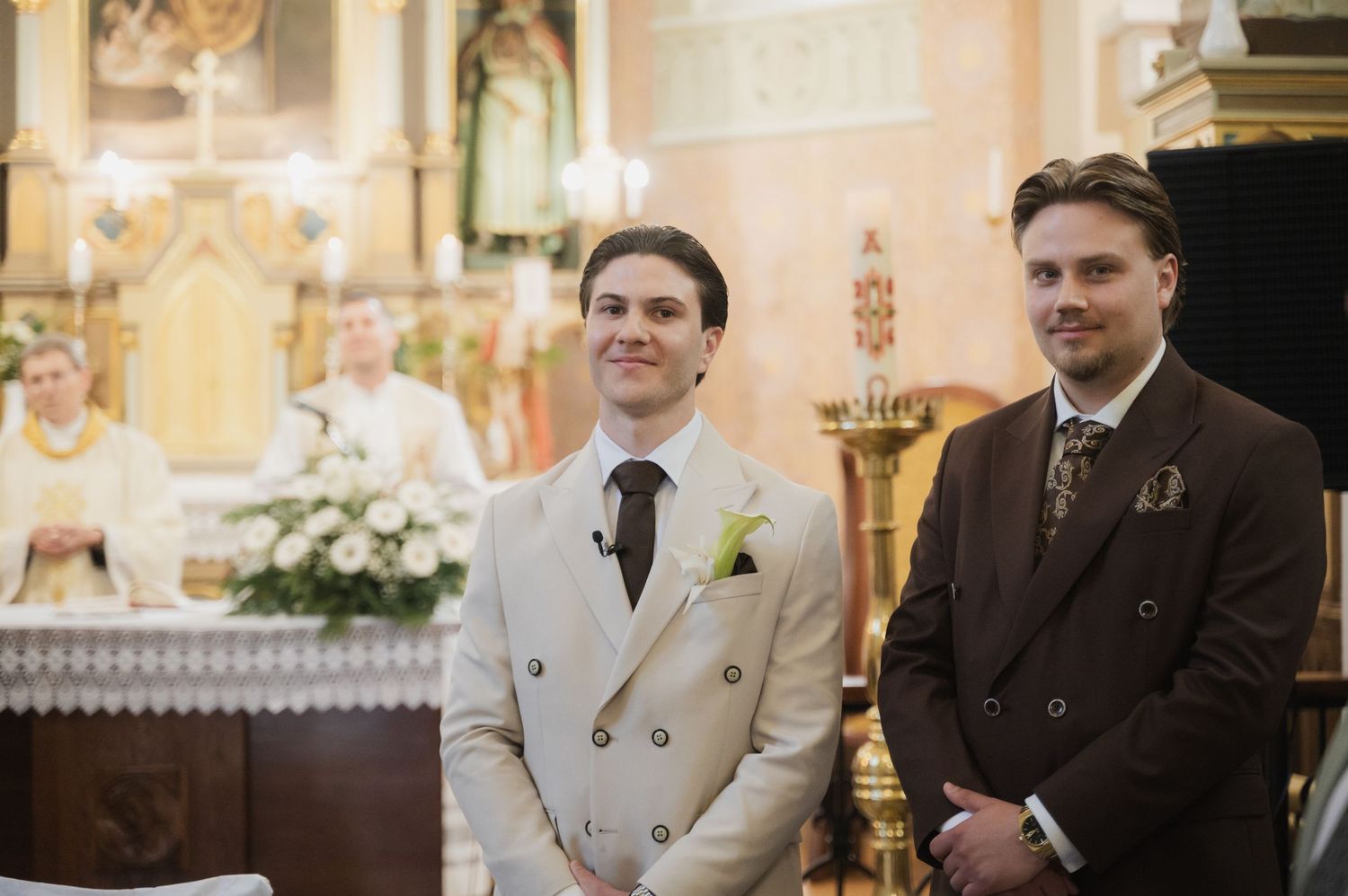 Groom in cream double-breasted suit stands with best man in brown suit at church wedding altar ceremony.