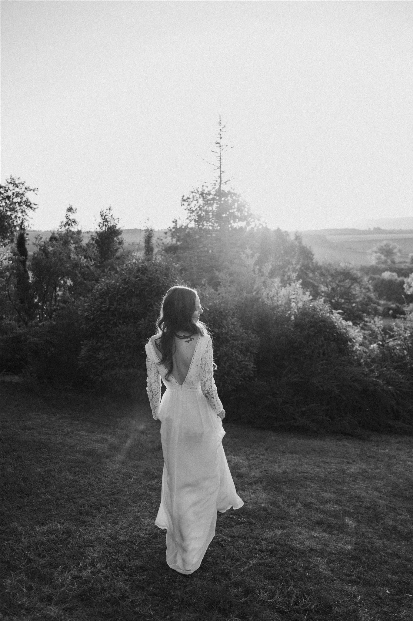 A silhouette of a bride in a flowing dress stands in a garden at sunset.