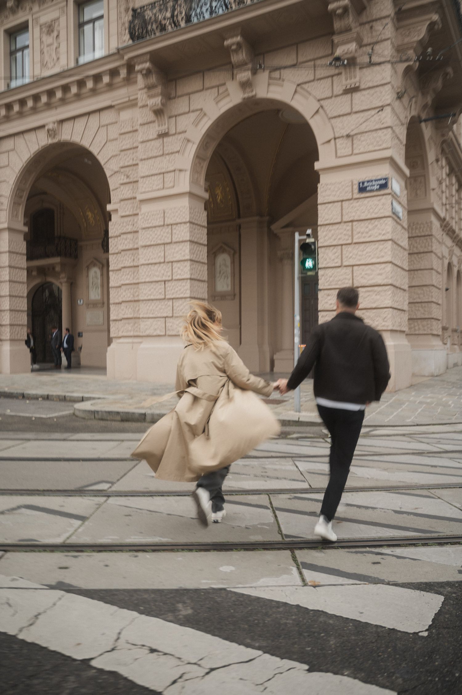 A person in a beige coat falls while walking on stone pavement next to a historic building facade.