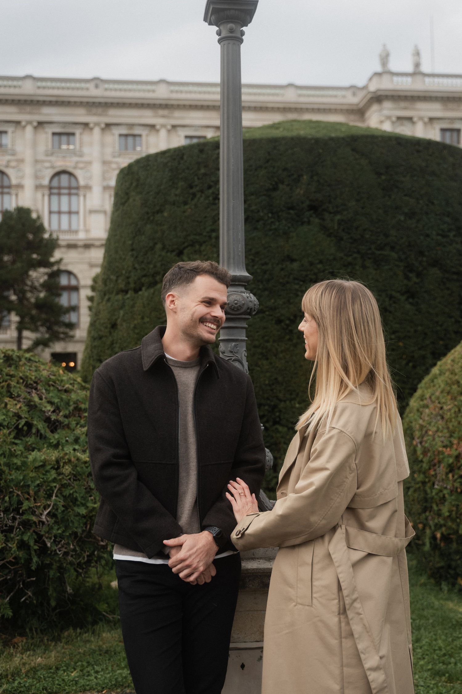 A couple in casual attire shares a joyful moment together while standing in a garden near historic architecture.