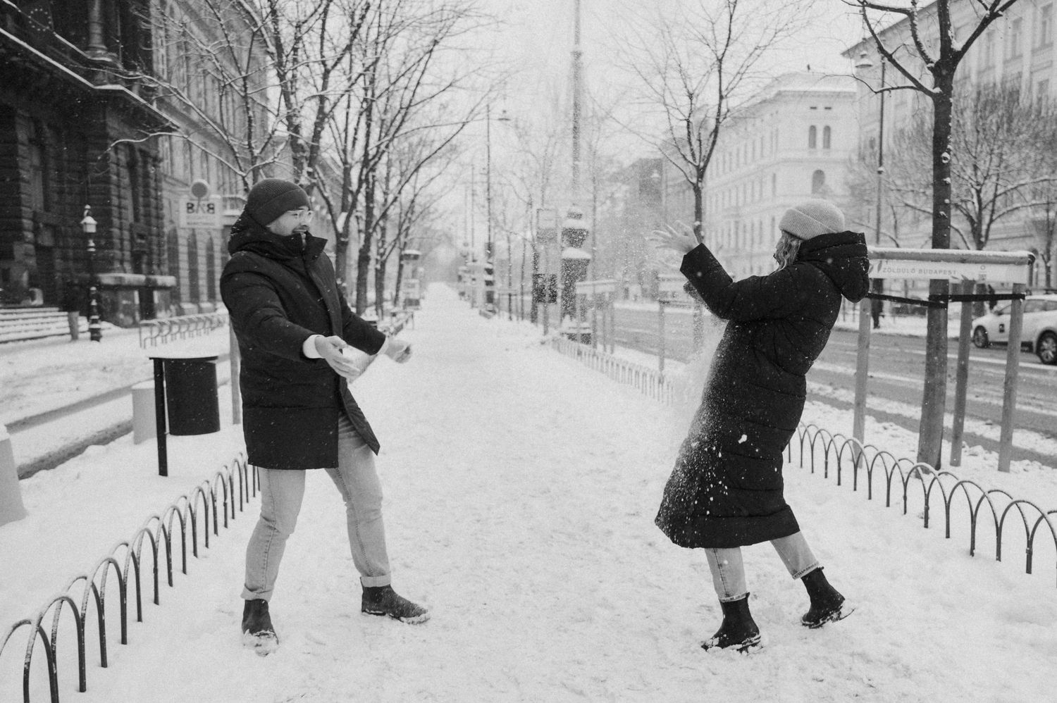 Two people playing and posing dramatically in the snow on a city sidewalk.