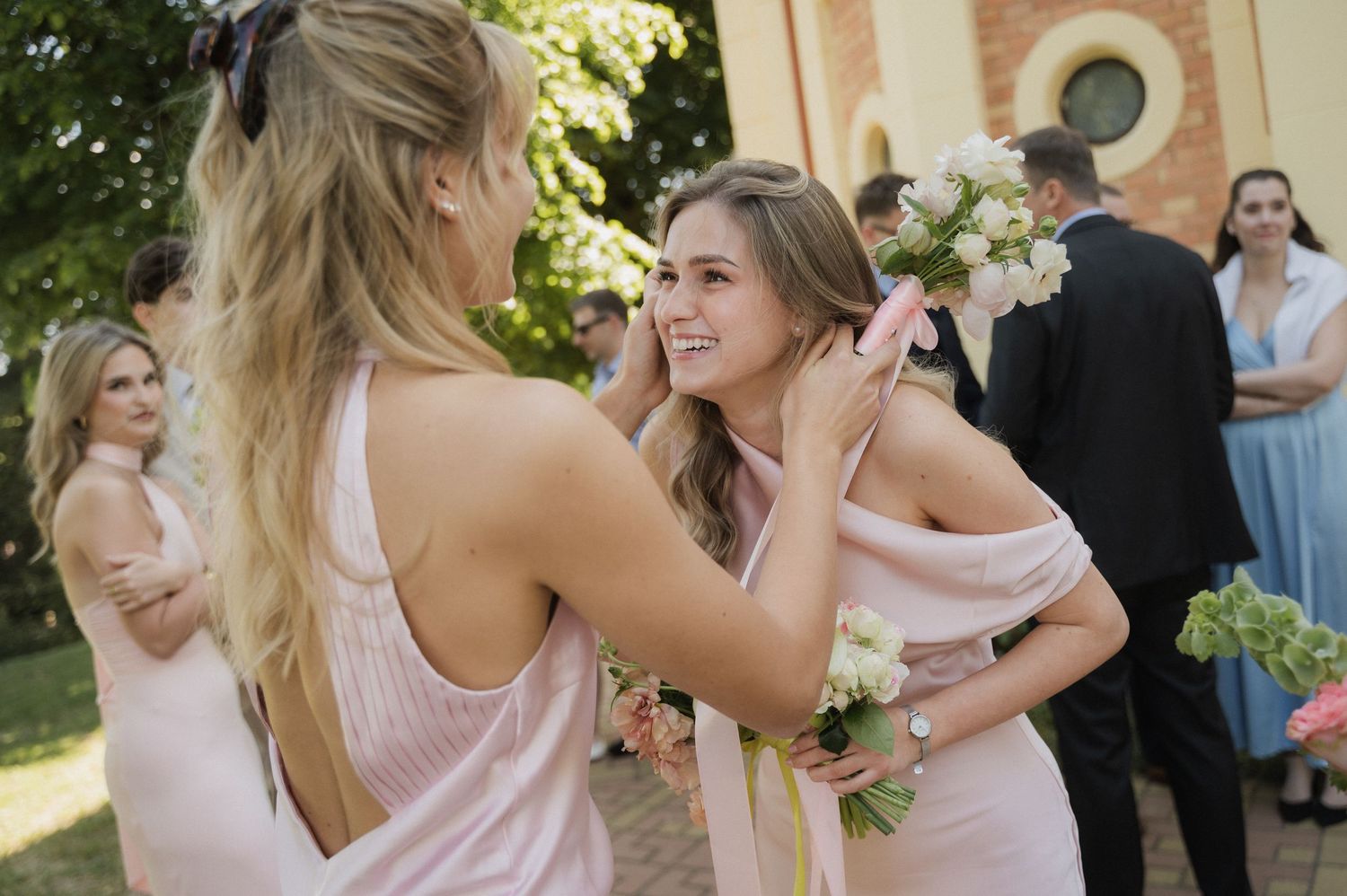 Bridesmaids in blush pink dresses laughing and embracing outside a church holding bouquets.