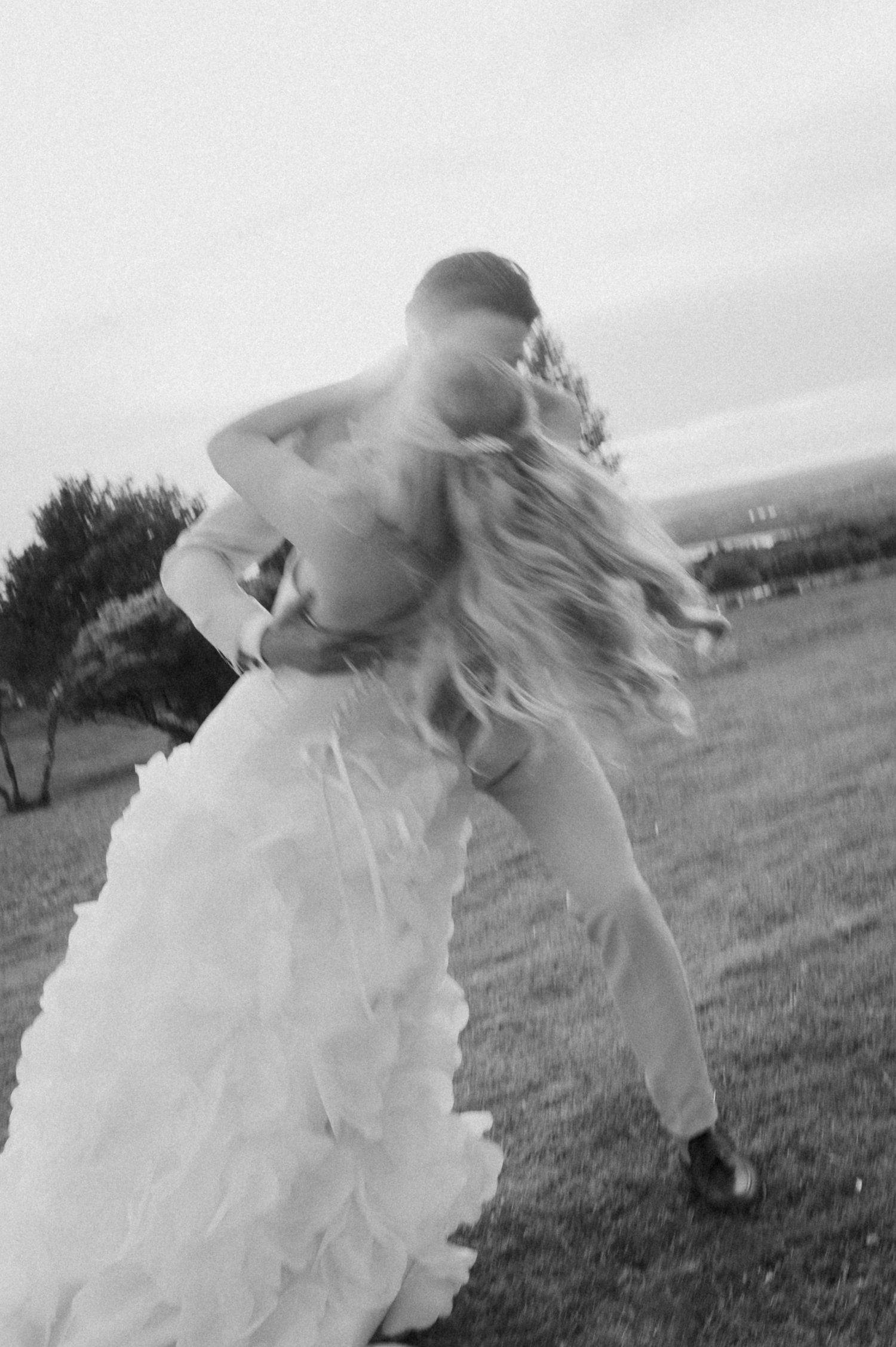 Black and white photo of bride and groom embracing outdoors, brides ruffled gown flowing in the wind.
