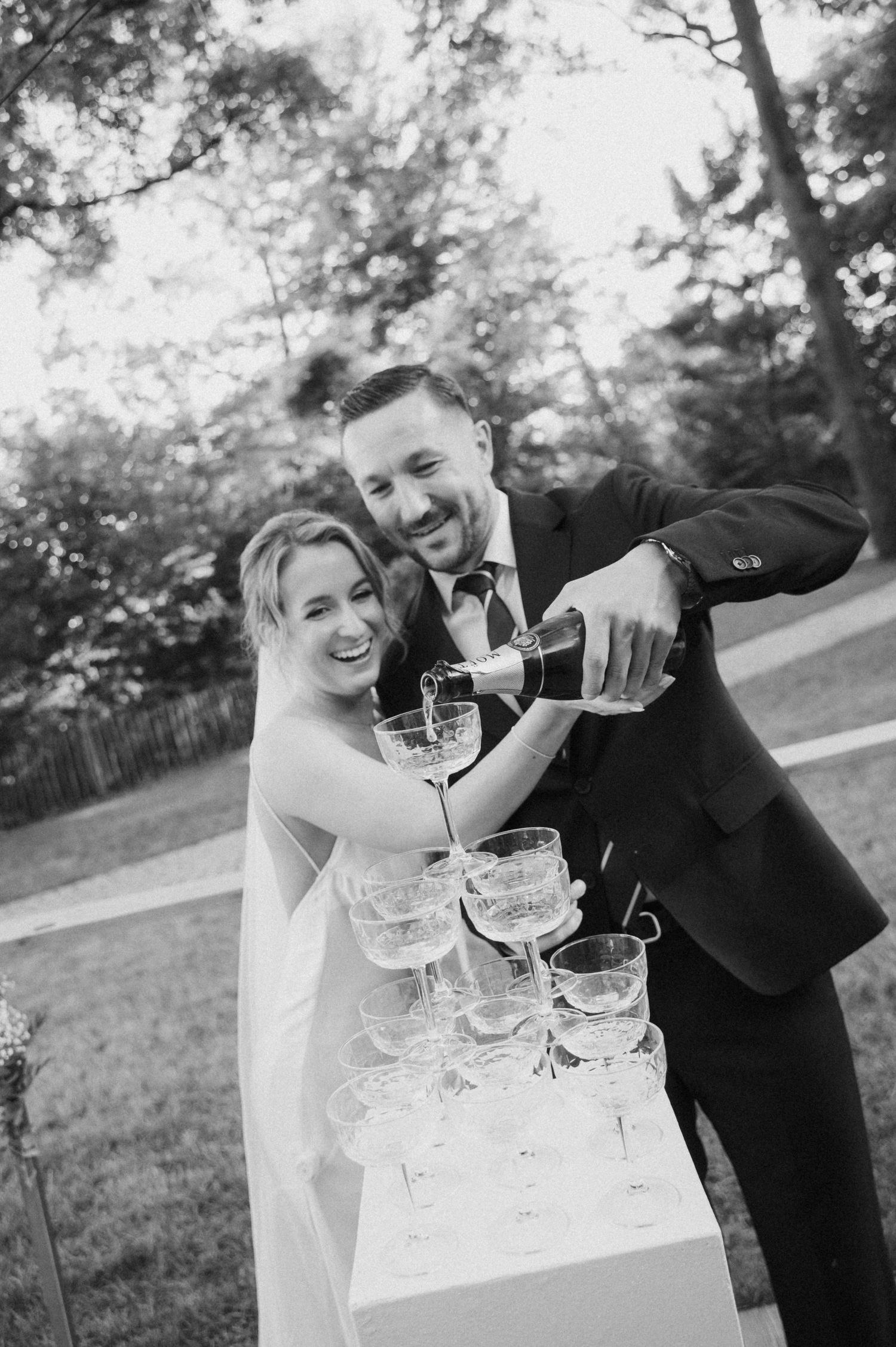 Smiling bride and groom pour champagne tower together at outdoor wedding reception in black and white.