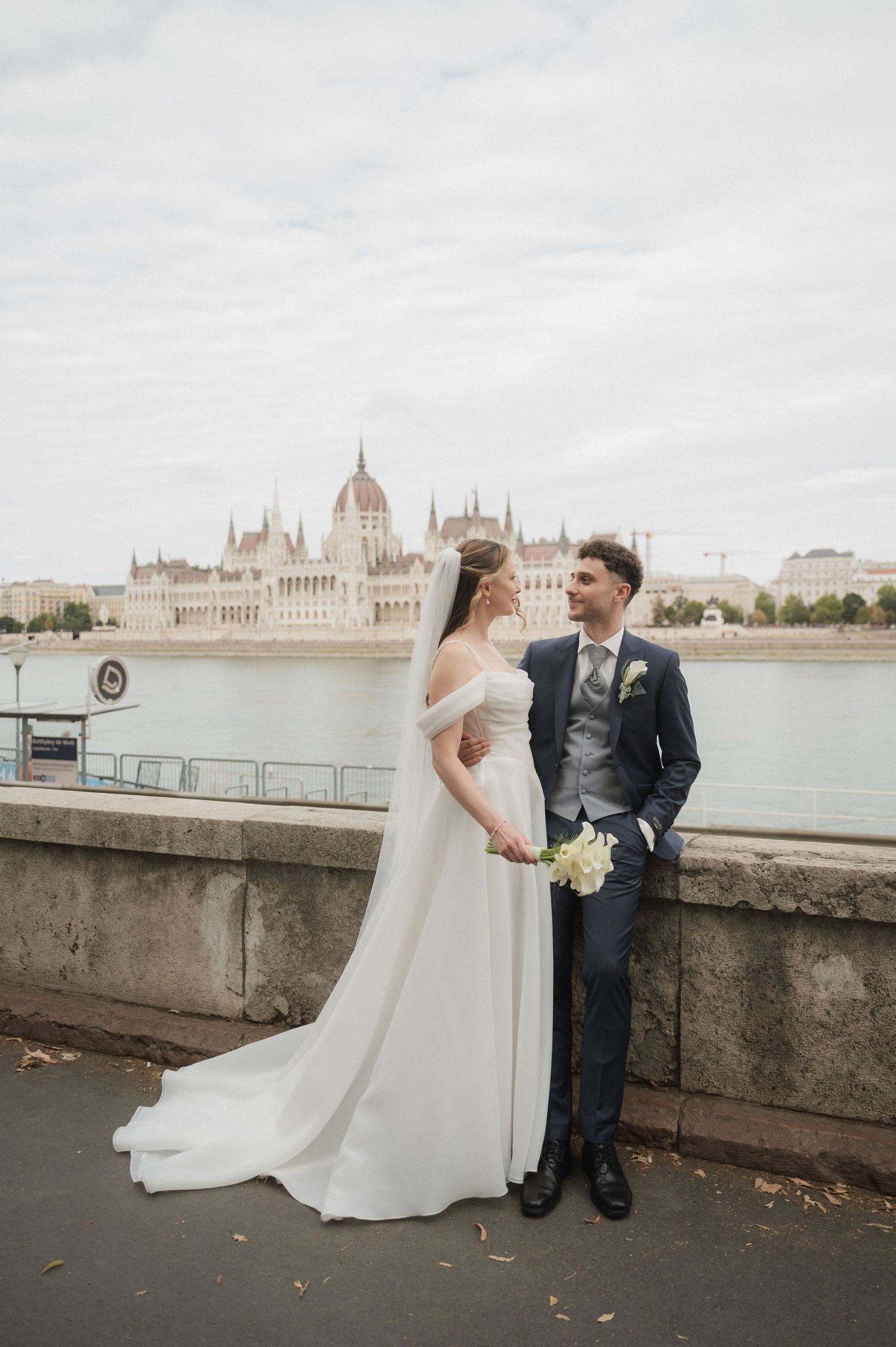 Bride and groom pose by riverside with Budapest Parliament building reflected in the Danube River behind them.