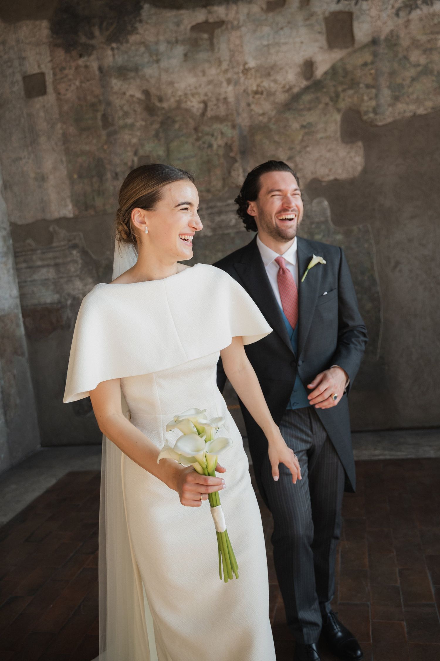 Joyful wedding scene with a bride in white holding calla lilies against weathered stone walls.