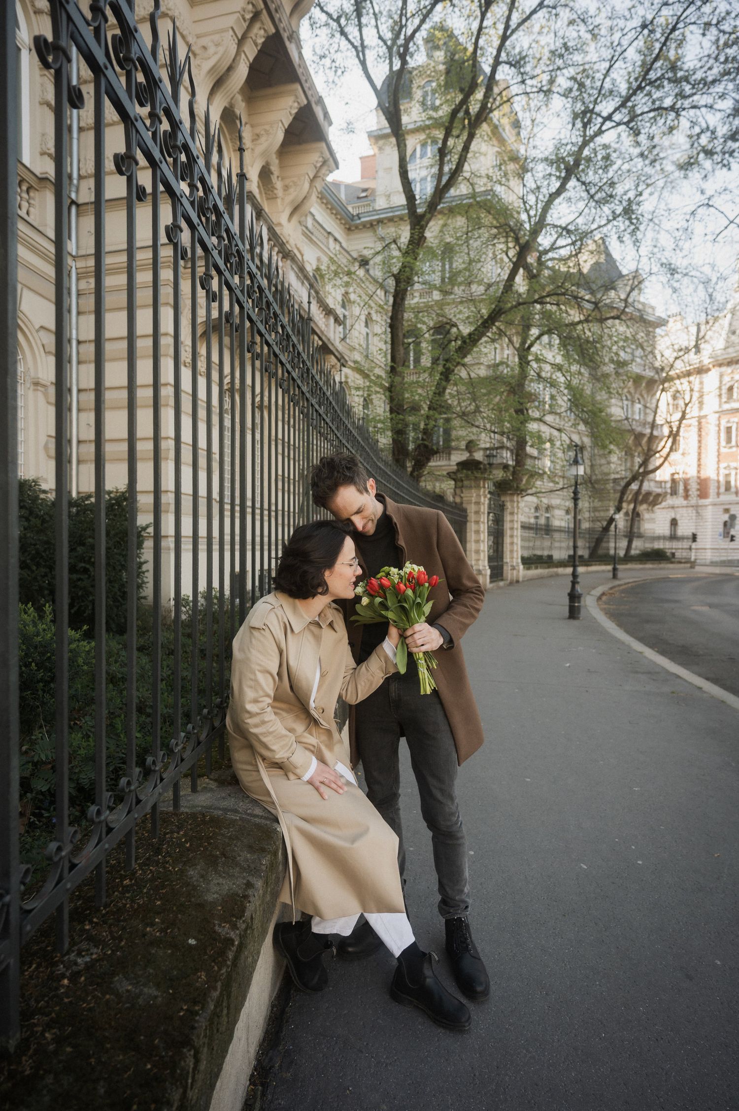 Two people share a romantic moment with flowers along an elegant colonnade walkway.