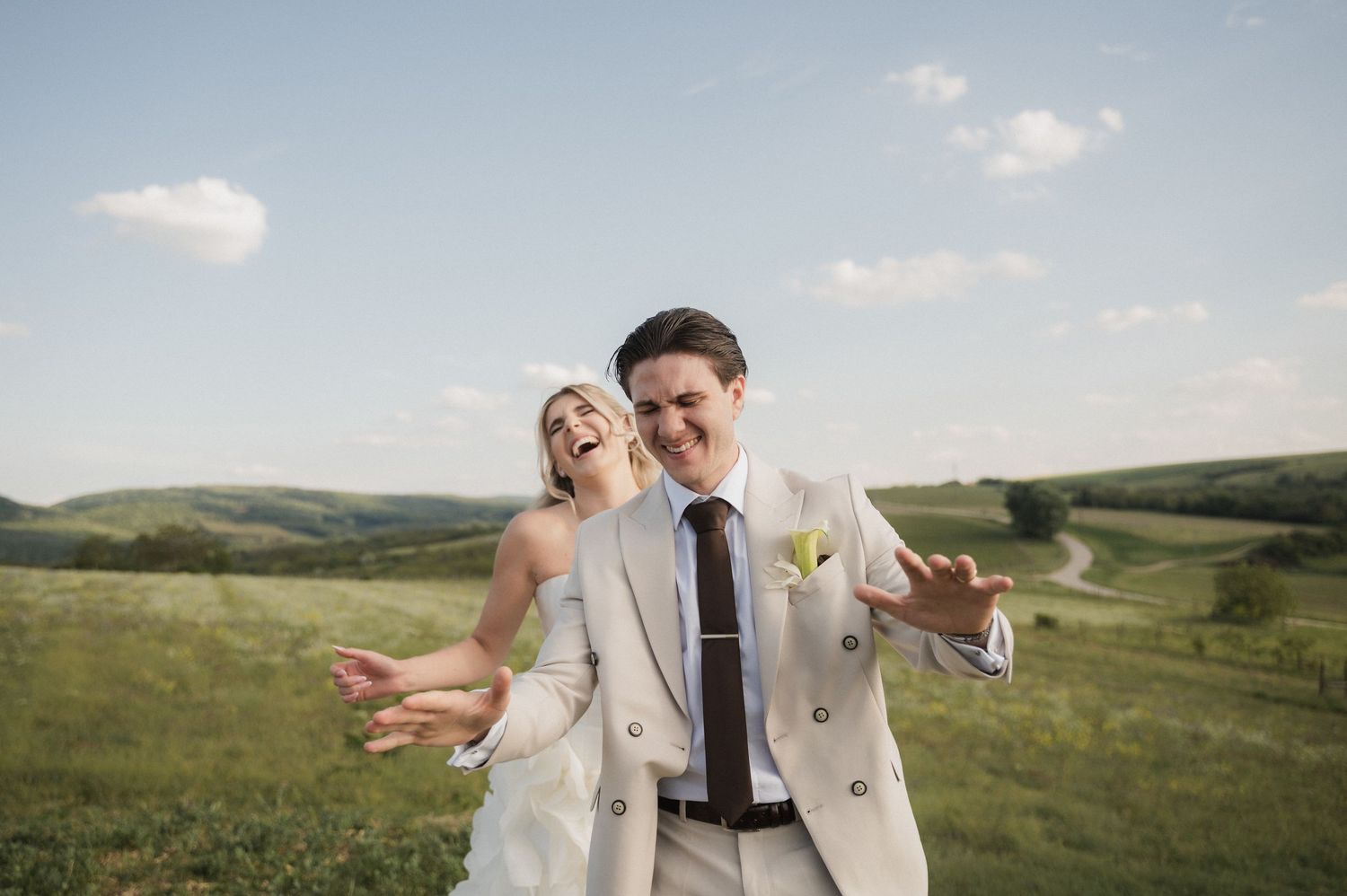 Joyful bride and groom laughing together in a scenic green countryside field on their wedding day.