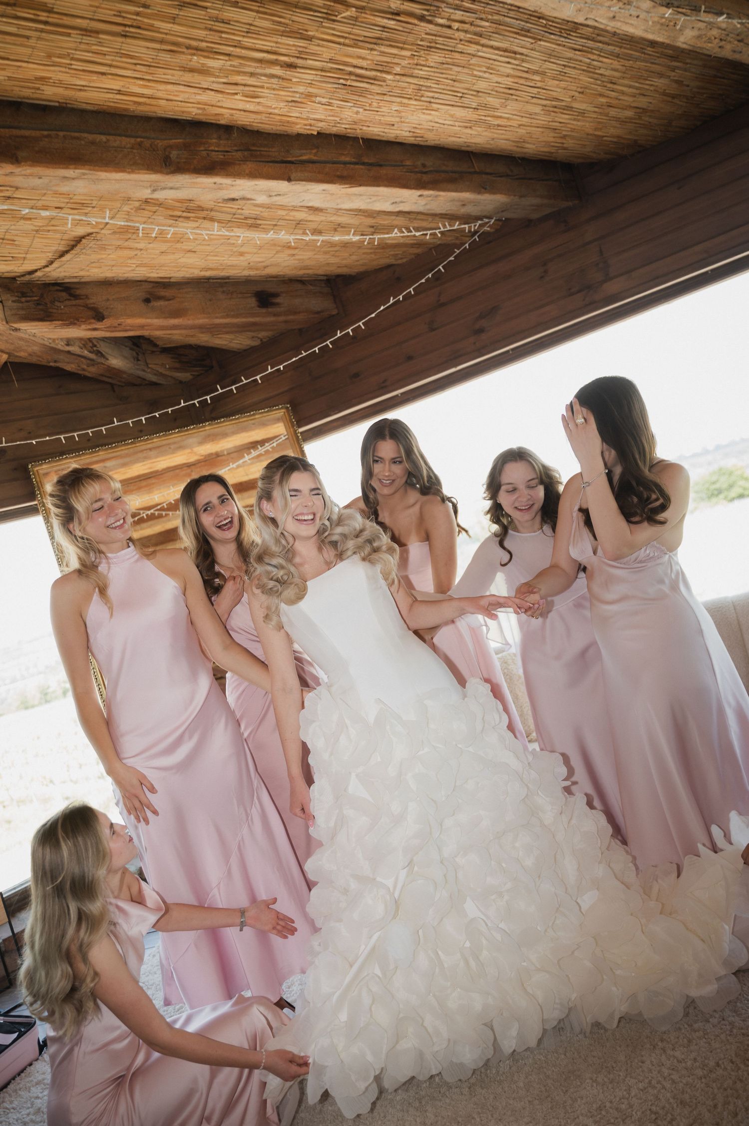 Bride in ruffled white gown laughing joyfully with six bridesmaids in blush pink dresses inside a rustic wooden venue.