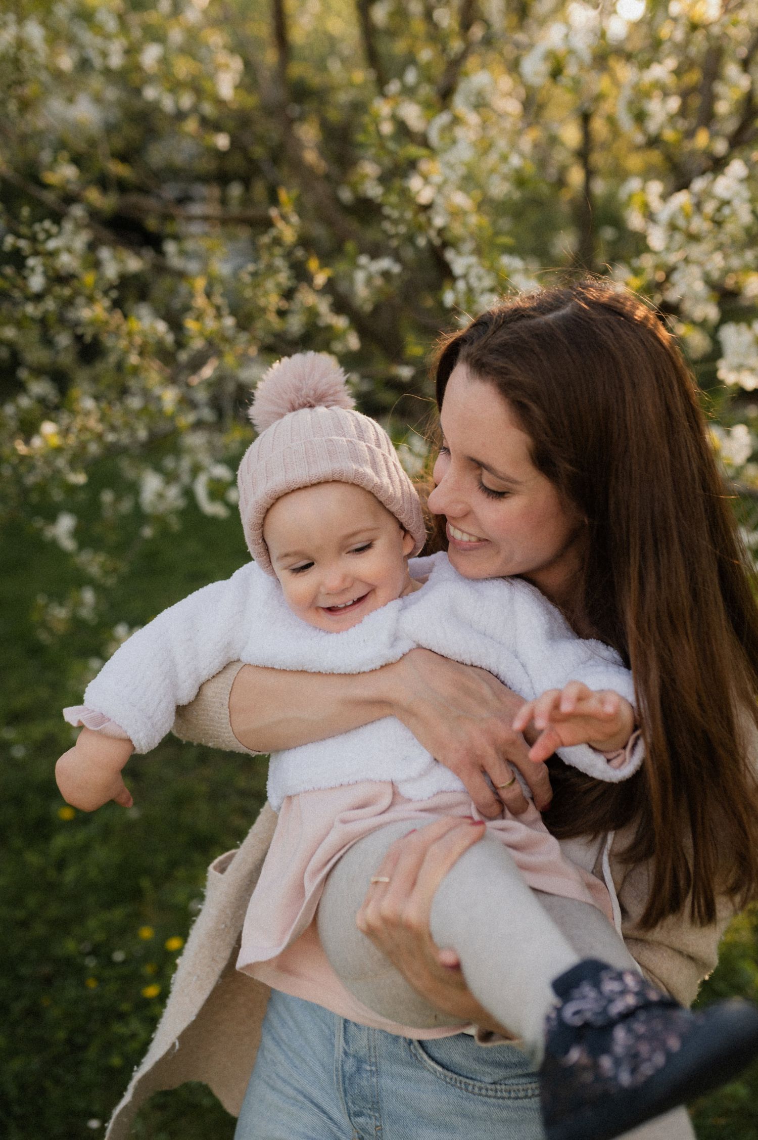 A smiling mother holds her happy baby in a pink knit hat among blooming spring trees outdoors.