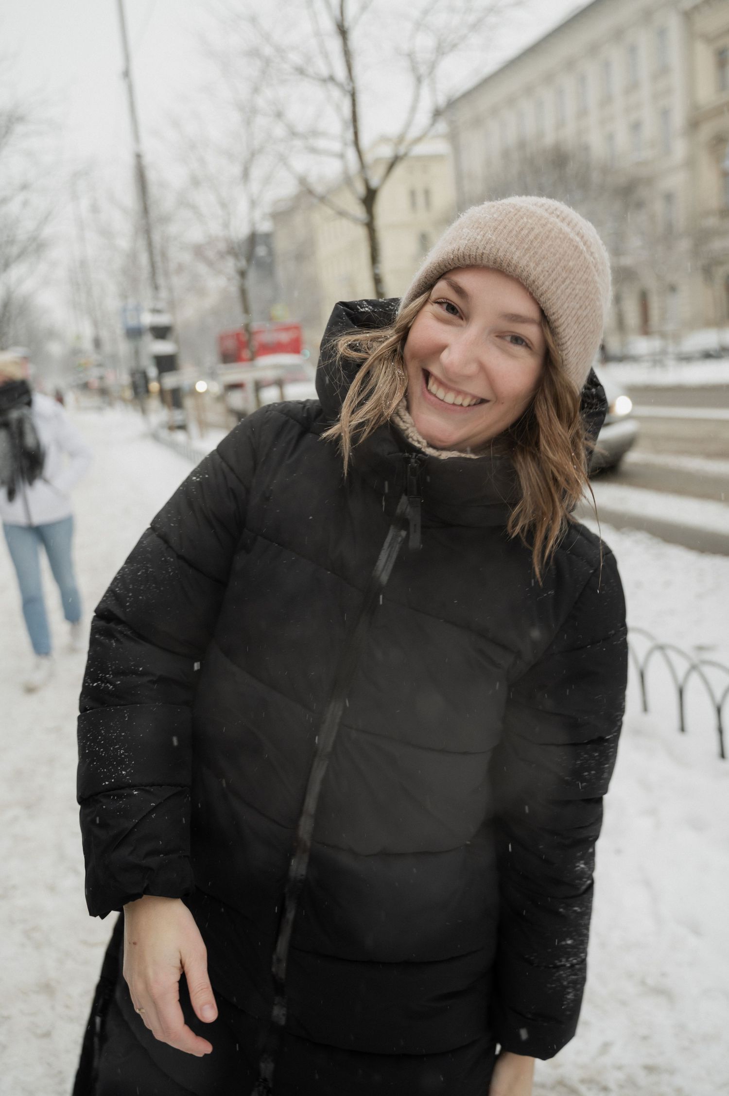 A joyful person in a winter coat and beanie smiles warmly on a snowy day.