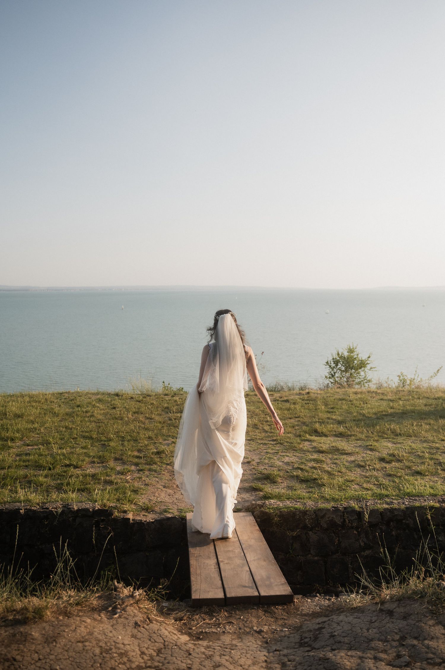 A figure in a flowing white dress walks along a wooden boardwalk path overlooking the ocean at sunset.