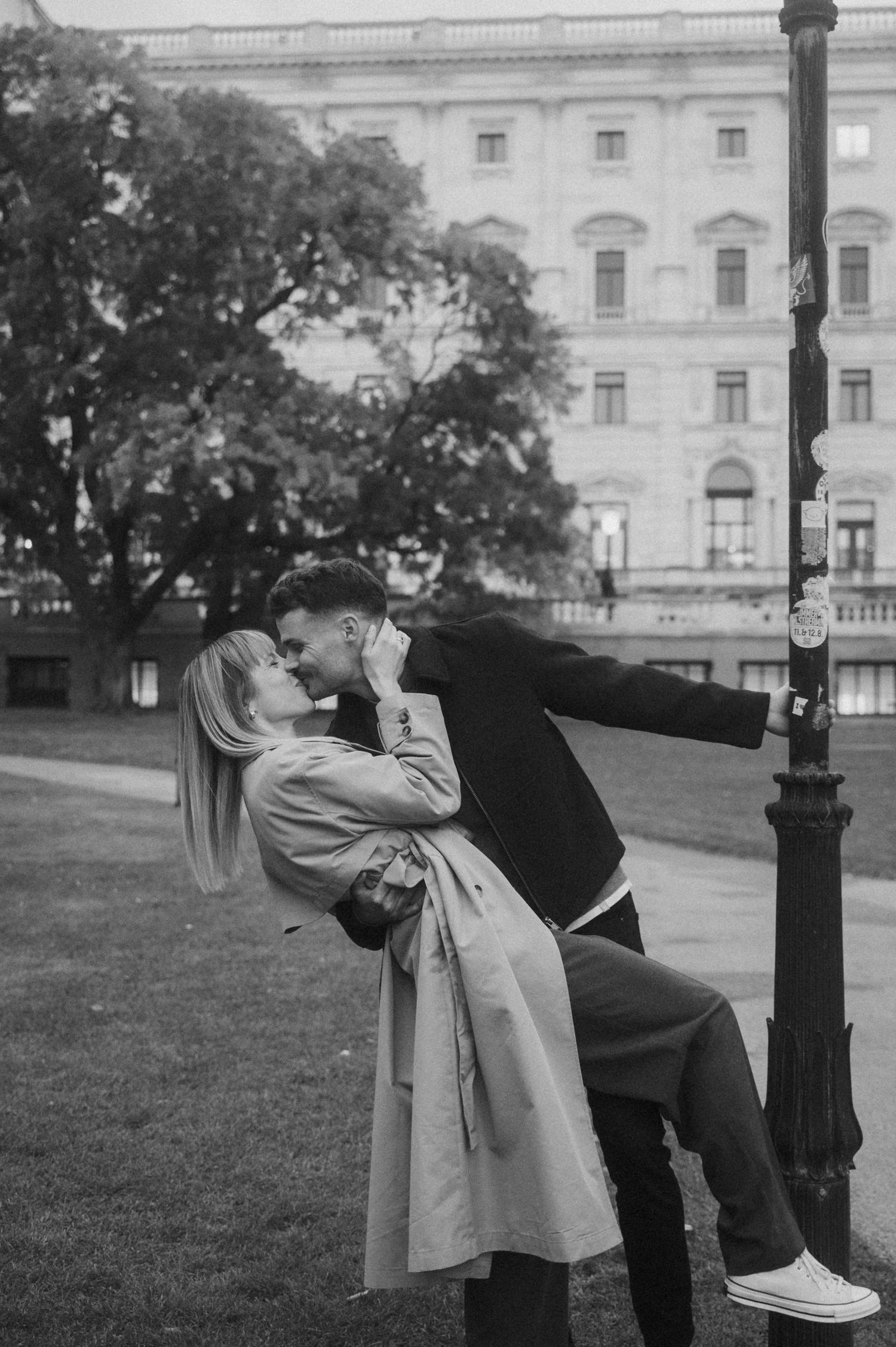 A couple in vintage attire shares a romantic kiss while leaning against a lamppost in front of a grand building.