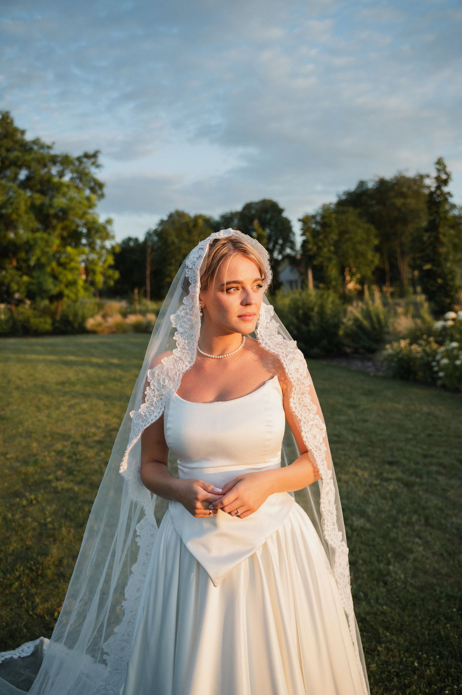 Bride in elegant white dress and veil poses in golden sunlight on grassy field.