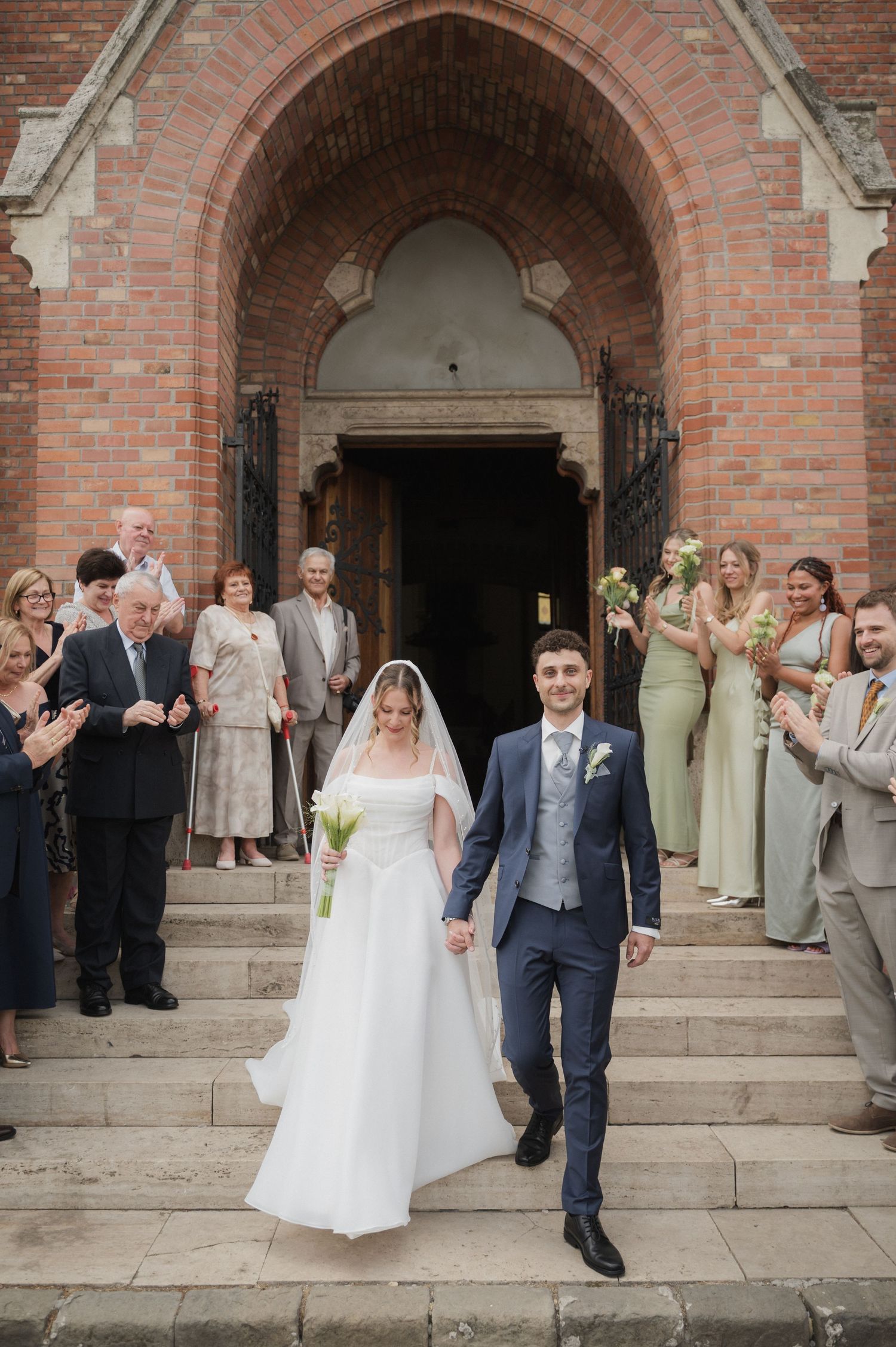 Newlyweds exit a red brick church greeted by applauding wedding guests on the steps.