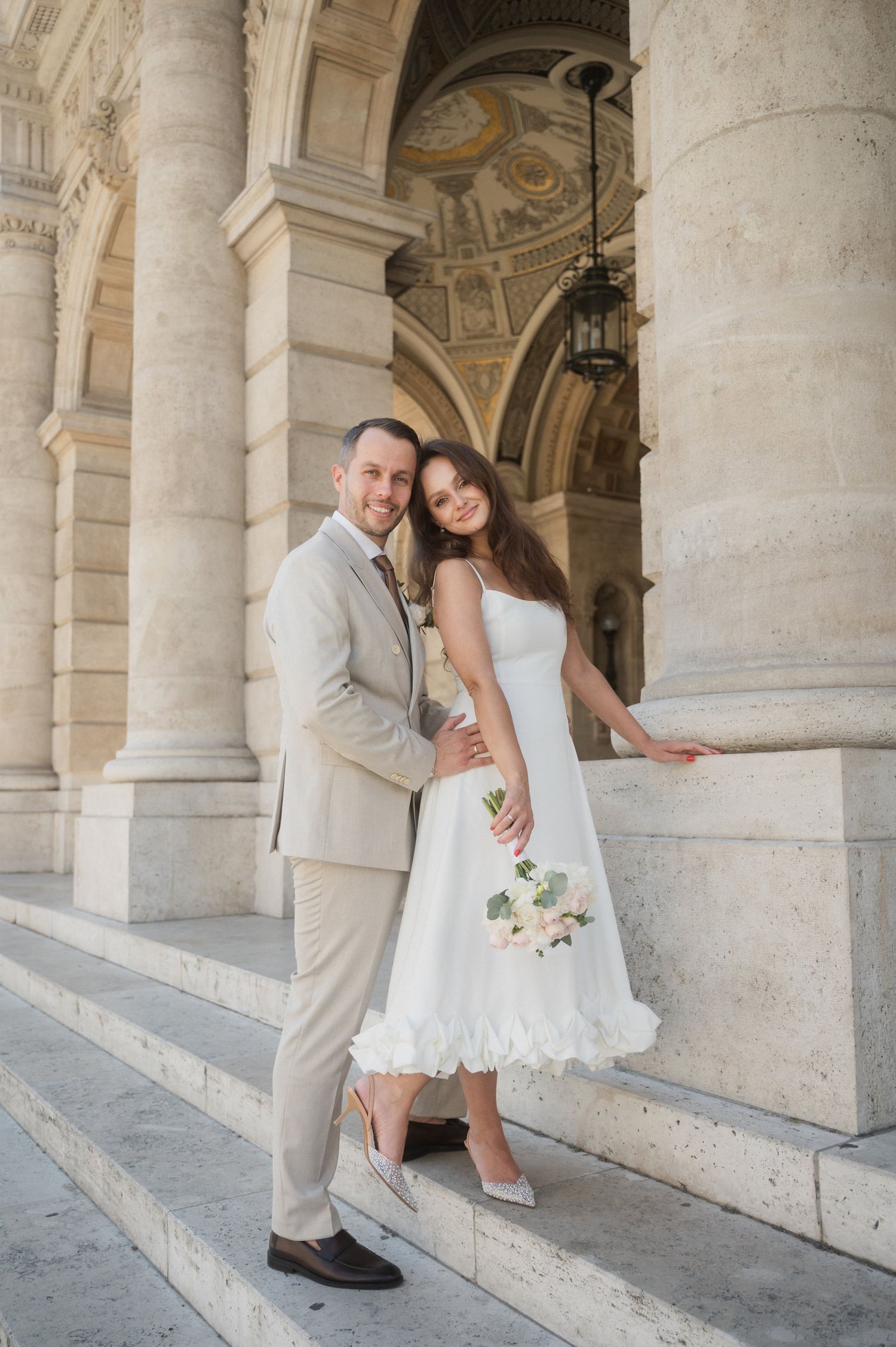 Romantic wedding couple poses playfully between elegant stone columns of a historic building.