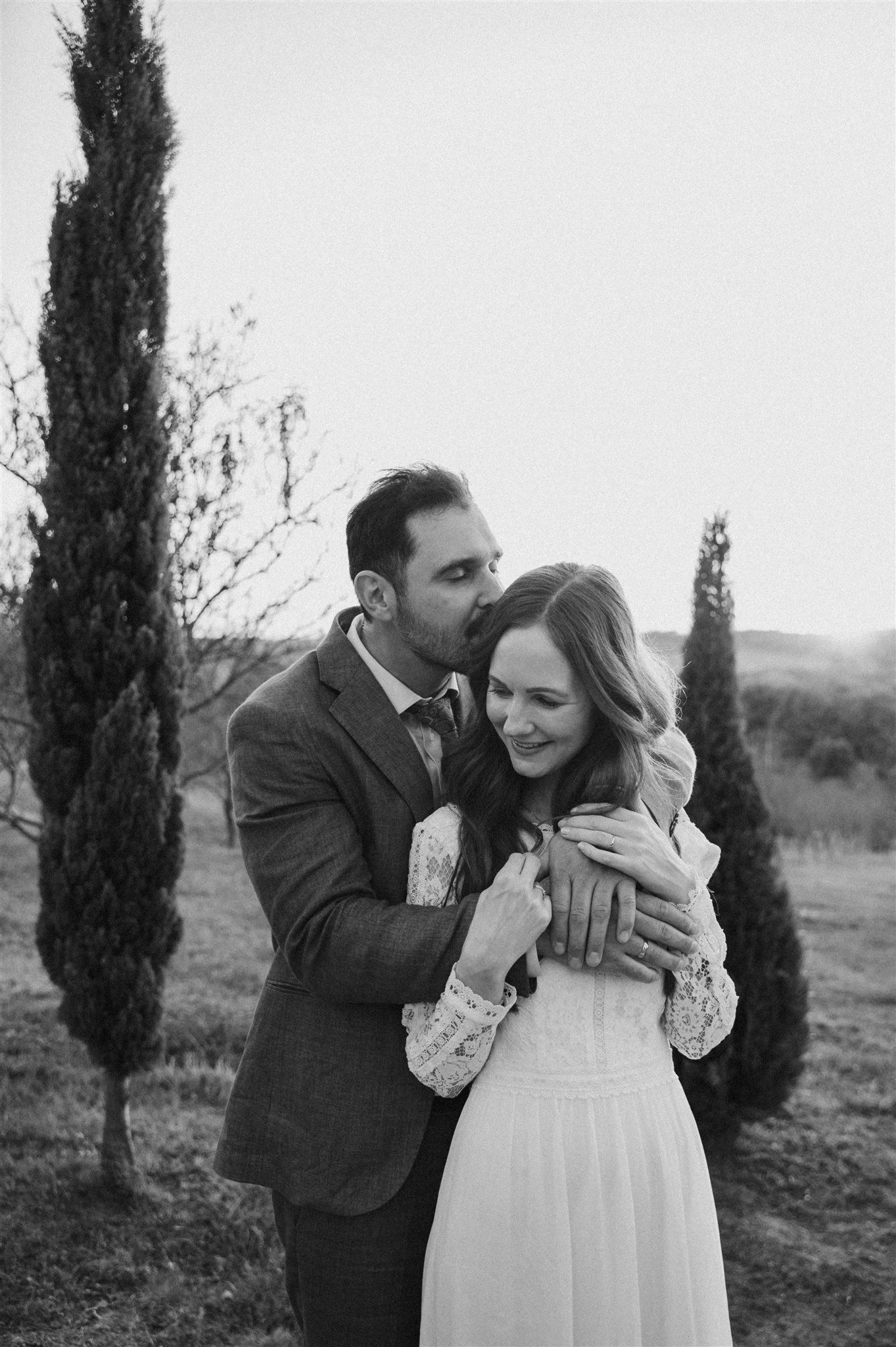 Black and white portrait of newlyweds embracing outdoors among cypress trees.