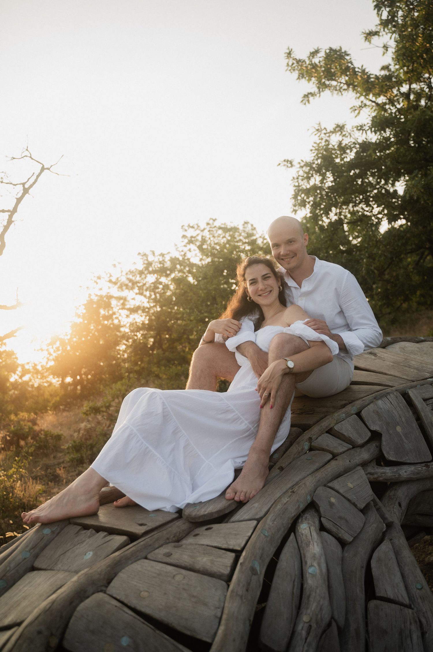 A couple in white clothing reclines on large rocks at sunset while sharing intimate moments in a romantic outdoor setting.