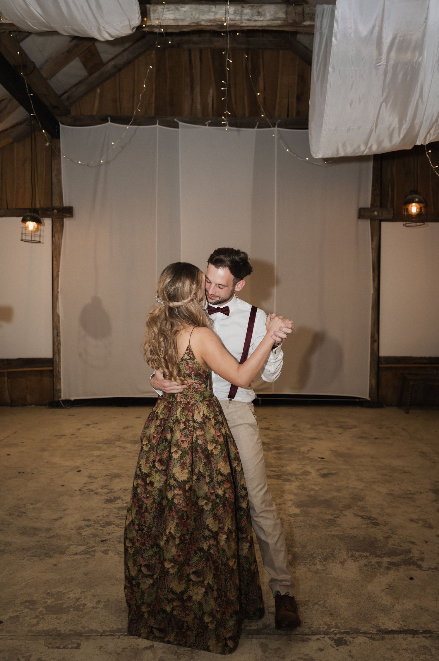 A couple dances together in a rustic barn venue with white drapes and wooden beams overhead.