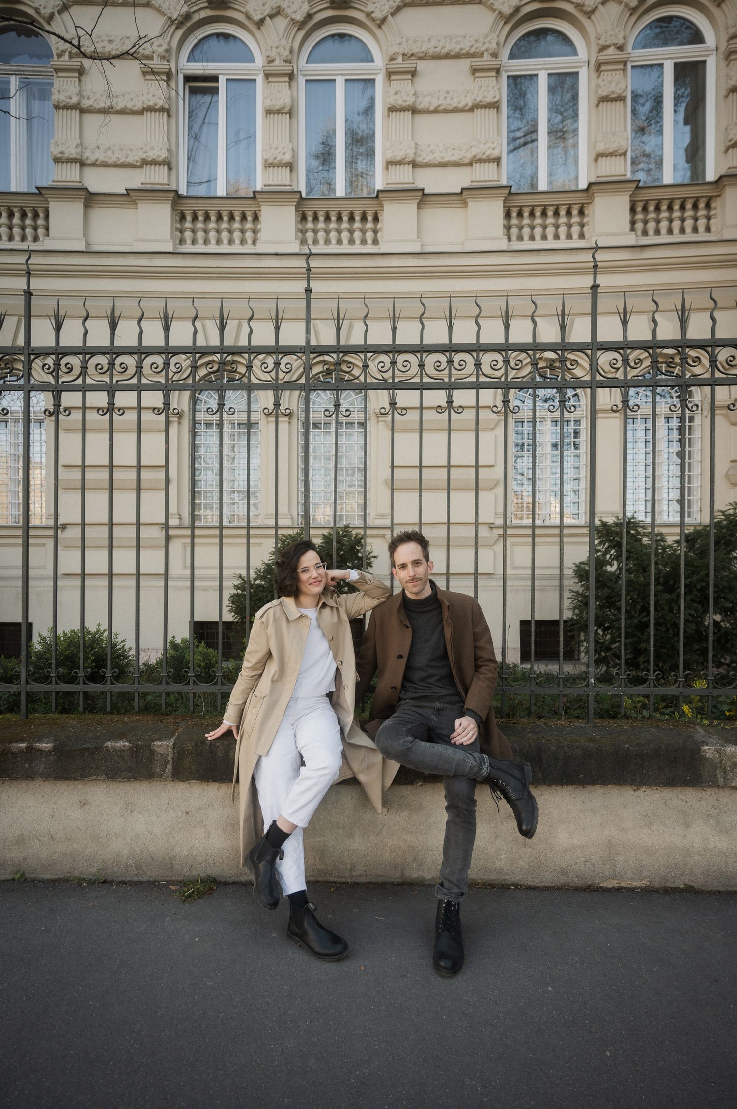 Two people sit casually on a stone wall outside an ornate European building with arched windows.