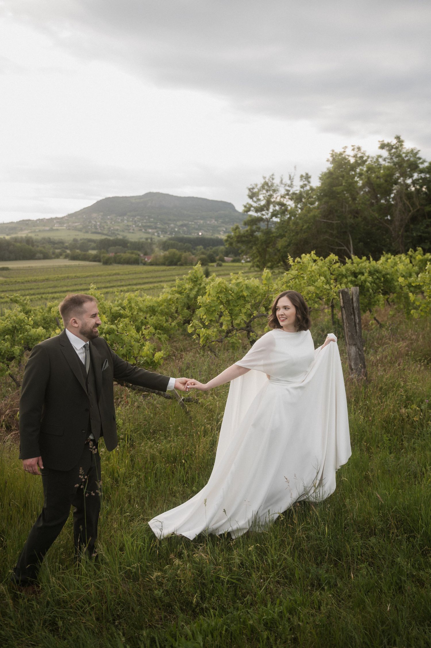 A couple in wedding attire dances playfully in a scenic vineyard with mountains visible in the background.