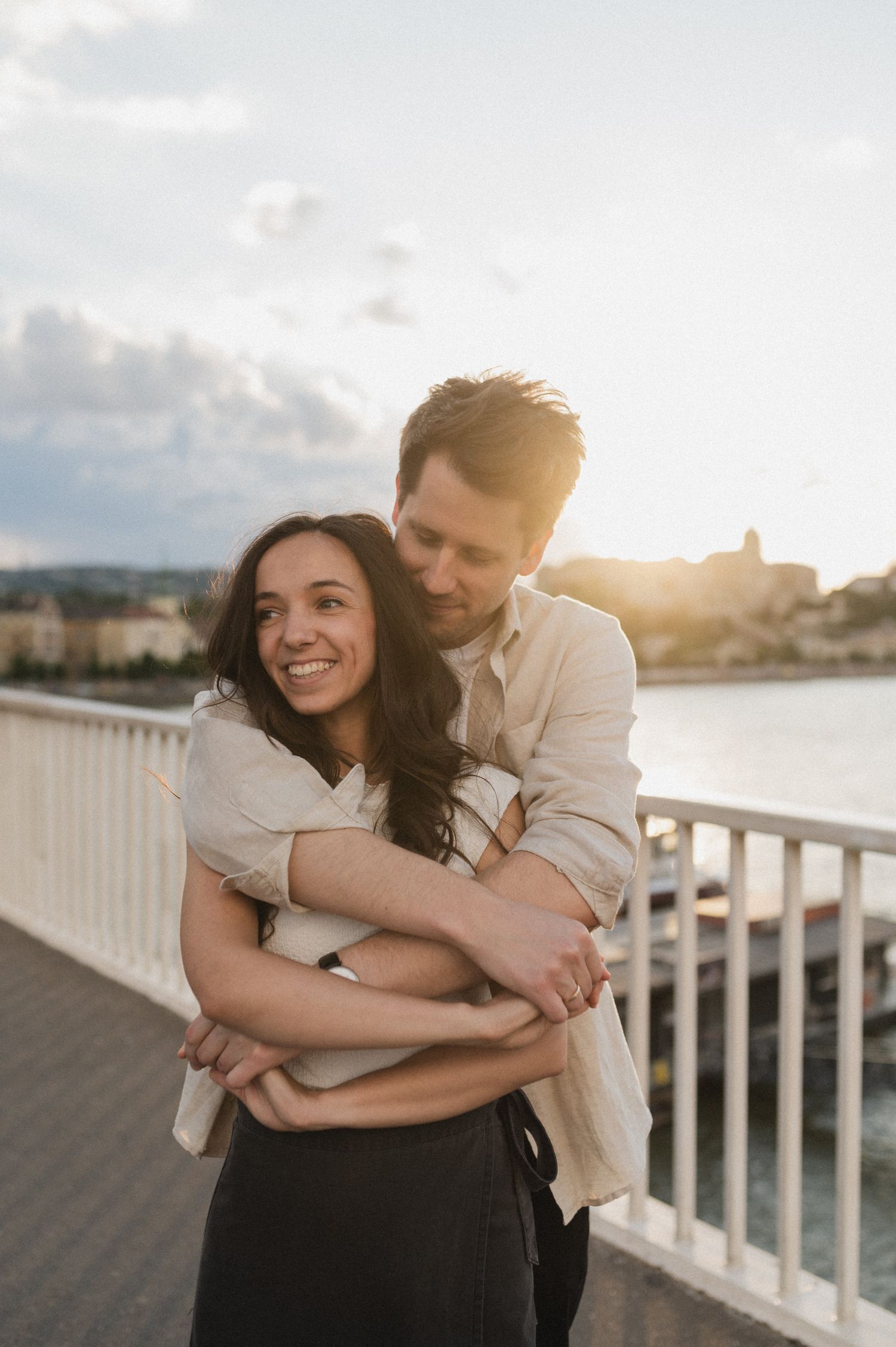 Romantic couple embraces on waterfront pier during golden sunset while leaning against white railing.