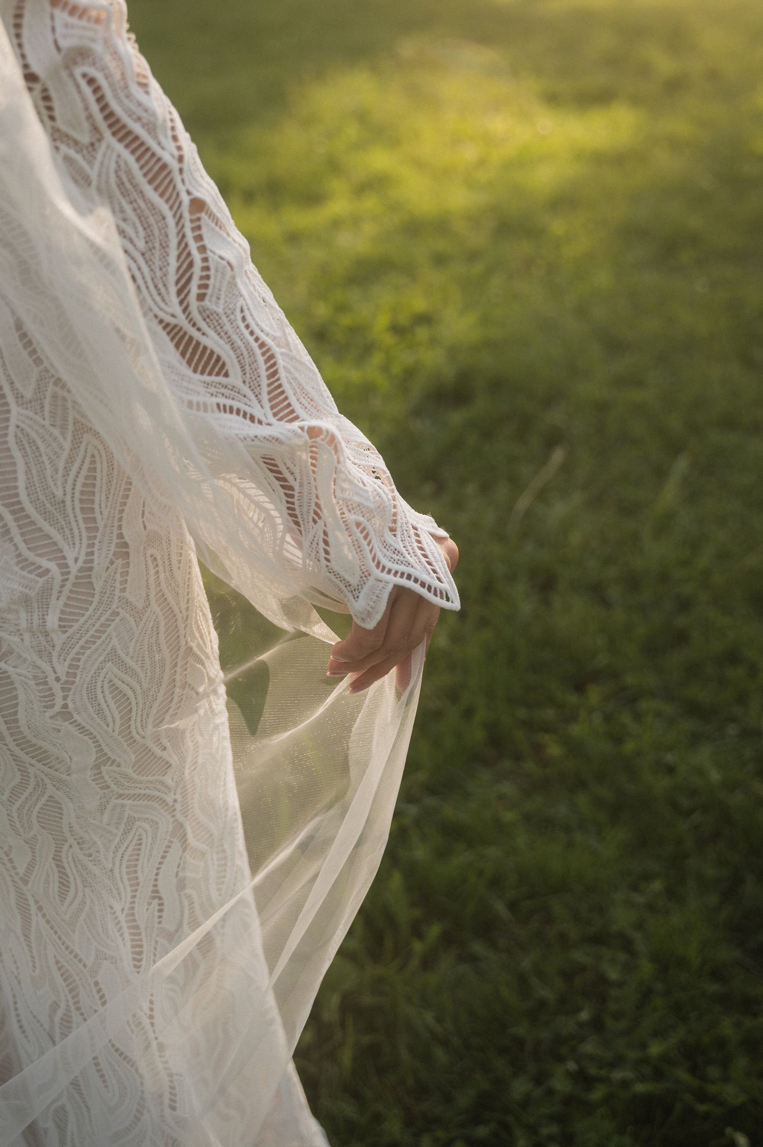 Close-up detail of a white lace wedding dress flowing in the breeze against a green grassy background.
