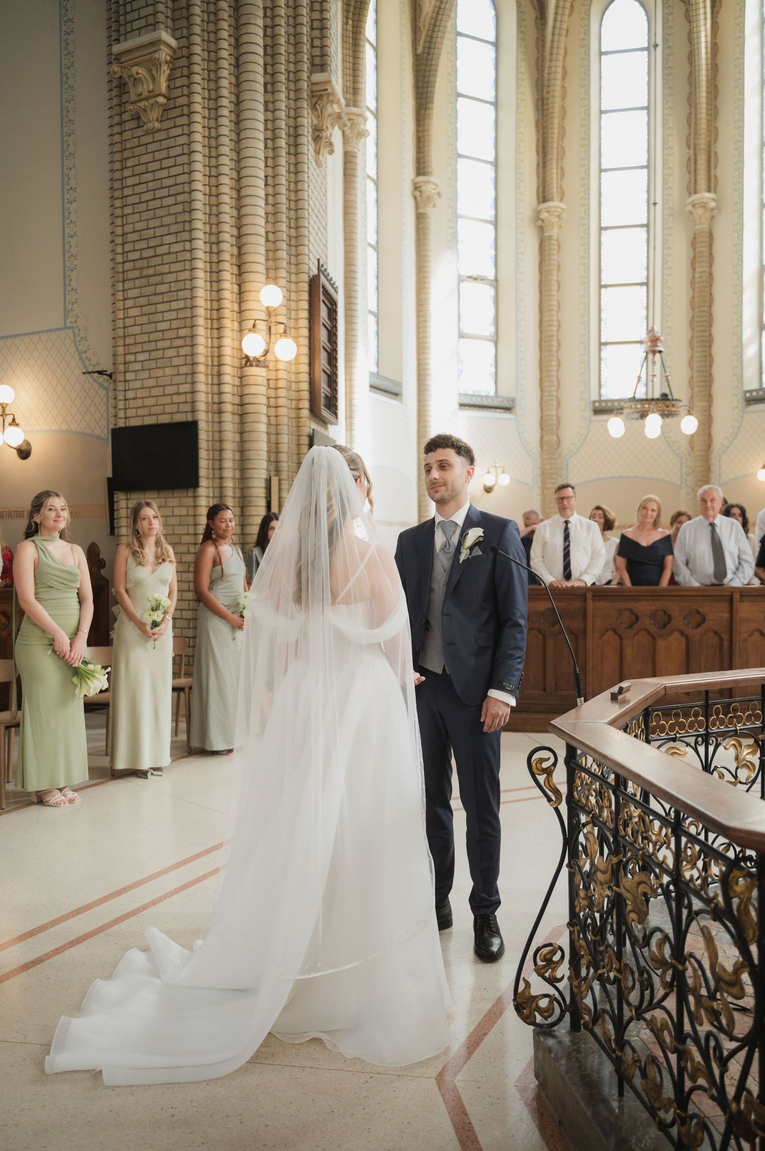 Bride and groom stand together at church altar during wedding ceremony with bridesmaids in sage green dresses nearby.