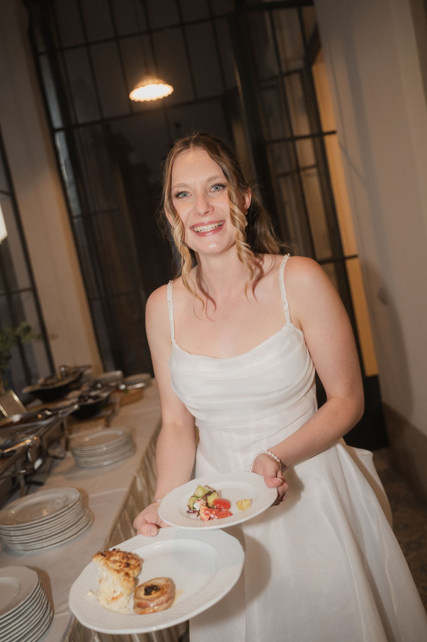 Smiling bride in white dress holding two plates of food at a wedding reception buffet.