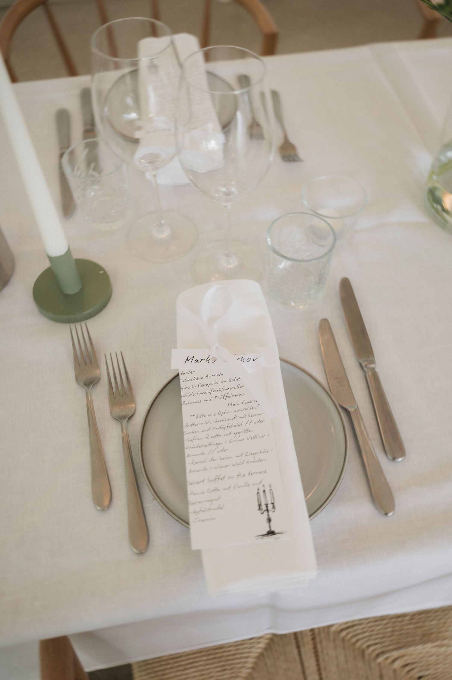 Elegant wedding place setting with menu card, silverware, glassware, and green taper candle on white linen.