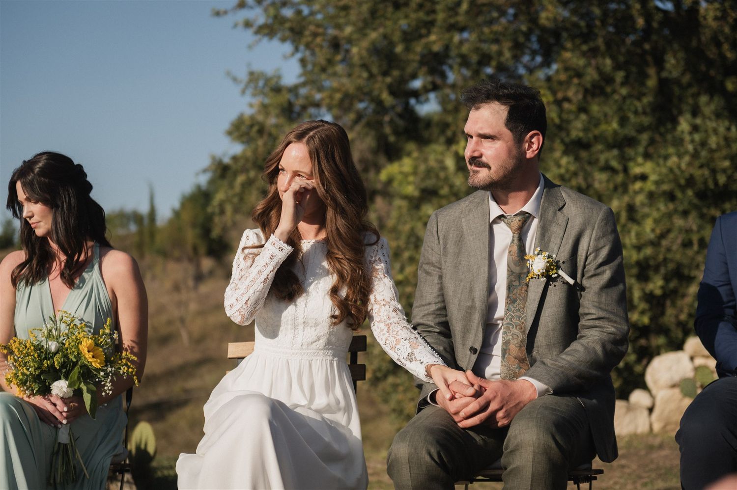 Wedding party members in sage green dresses and suits gather outdoors during golden hour.