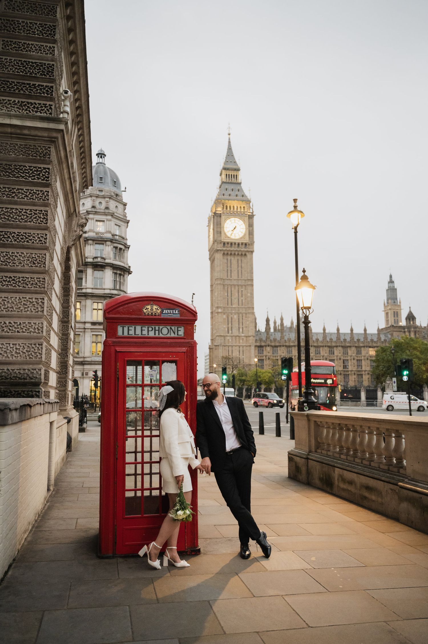 A stylish couple stands beside an iconic red phone box with Big Ben in the background.