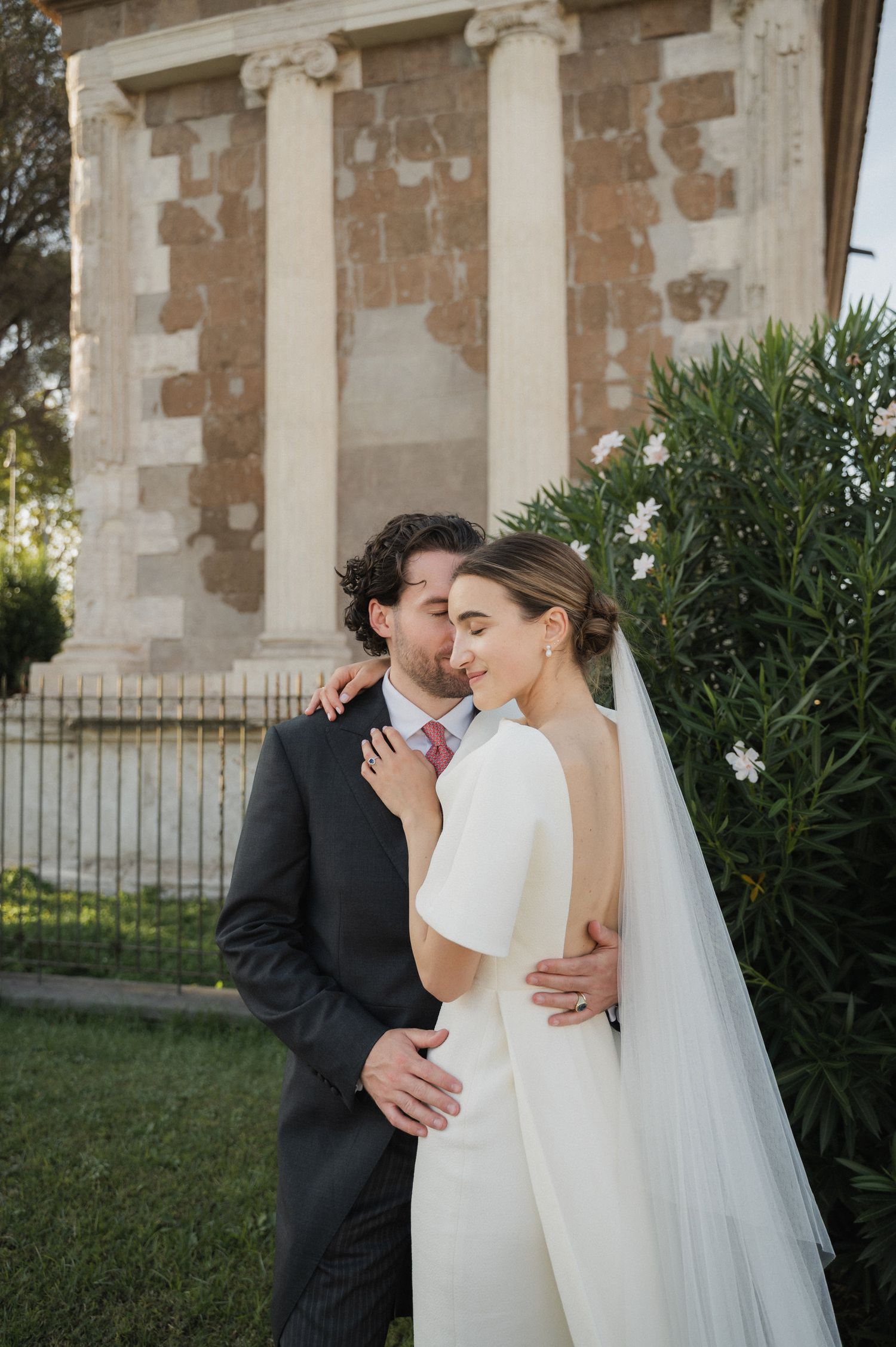 A romantic wedding moment captured against a stone wall with elegant columns and greenery.