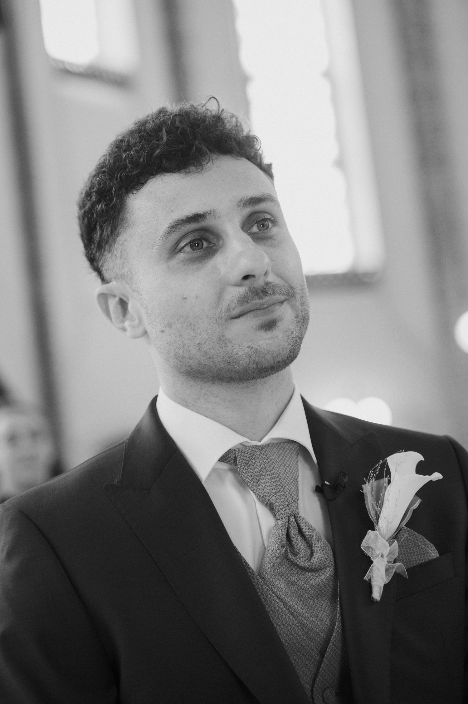 Black and white portrait of a groom in a suit with boutonniere, looking up emotionally during wedding ceremony.