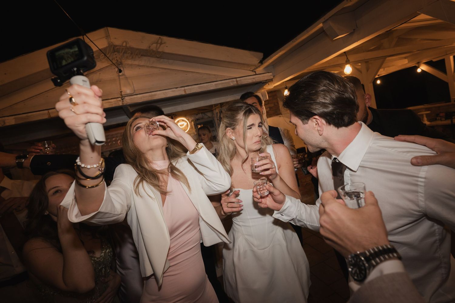 Wedding guests celebrate and take shots on lively dance floor during outdoor evening reception party.