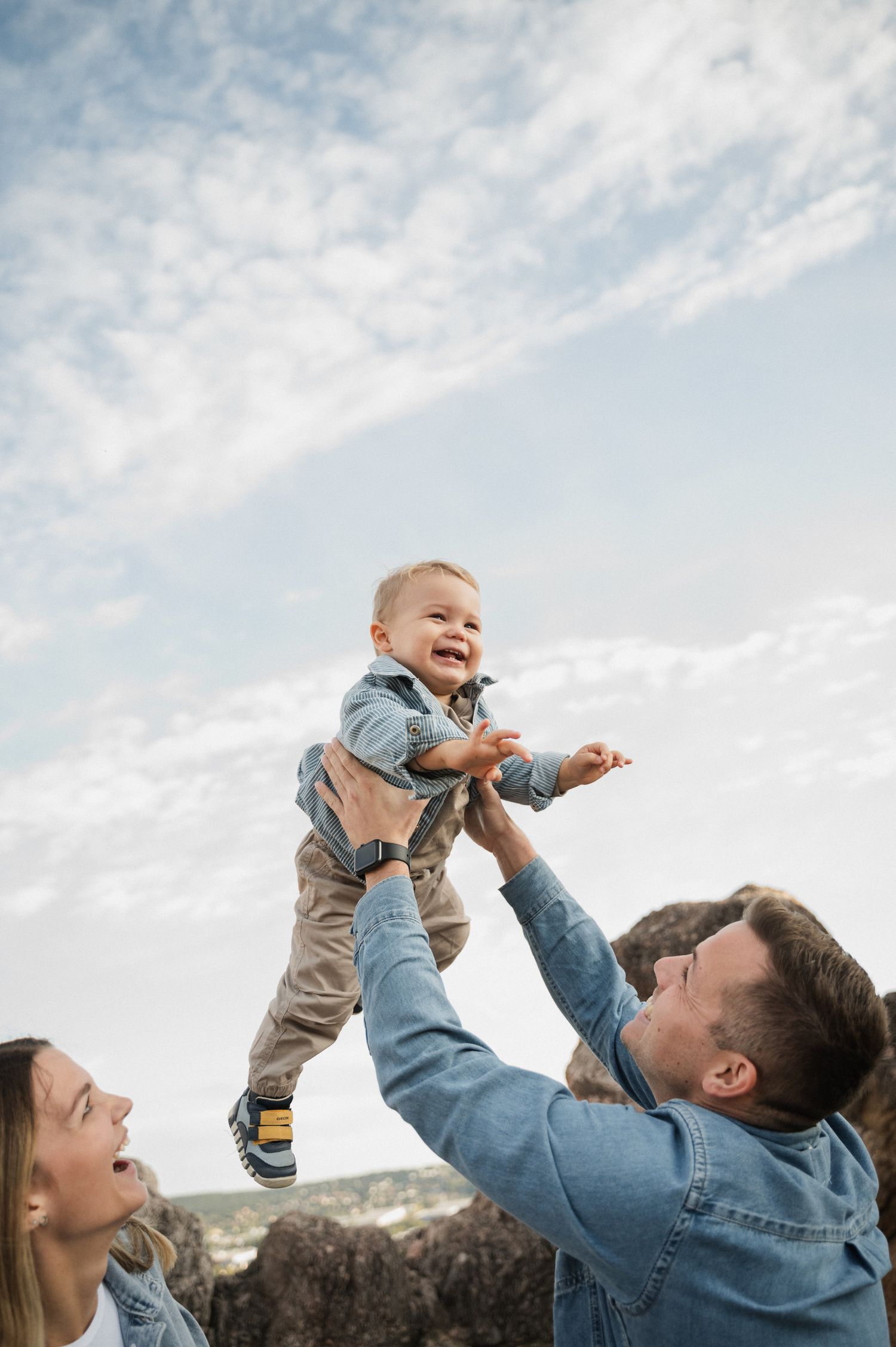A joyful family moment outdoors as a small child is lifted high into the air against a cloudy sky.