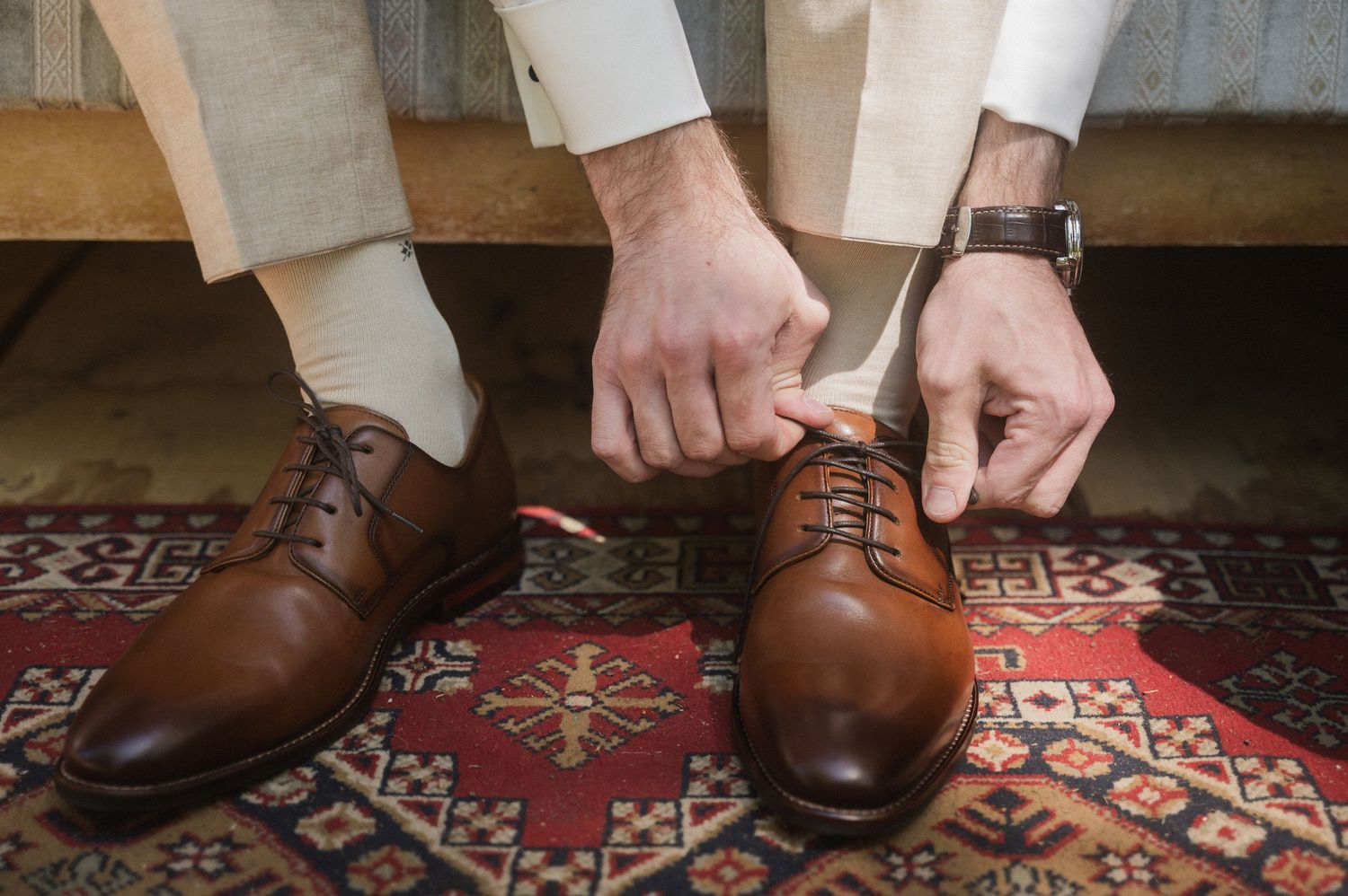 Close-up view of brown leather dress shoes being laced up on a decorative patterned rug.