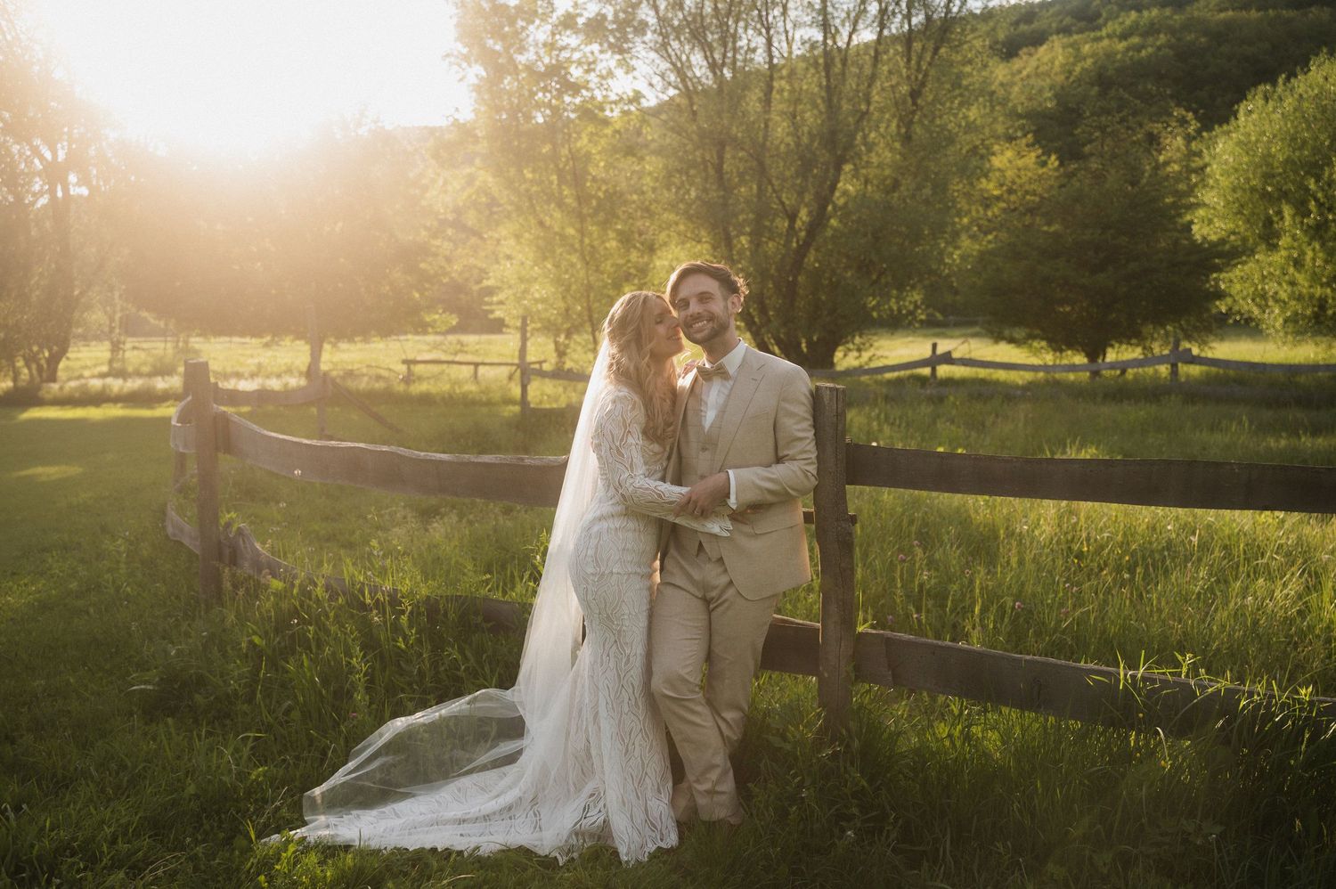 A couple embraces in a sunlit meadow during golden hour with rustic wooden fences in the background.