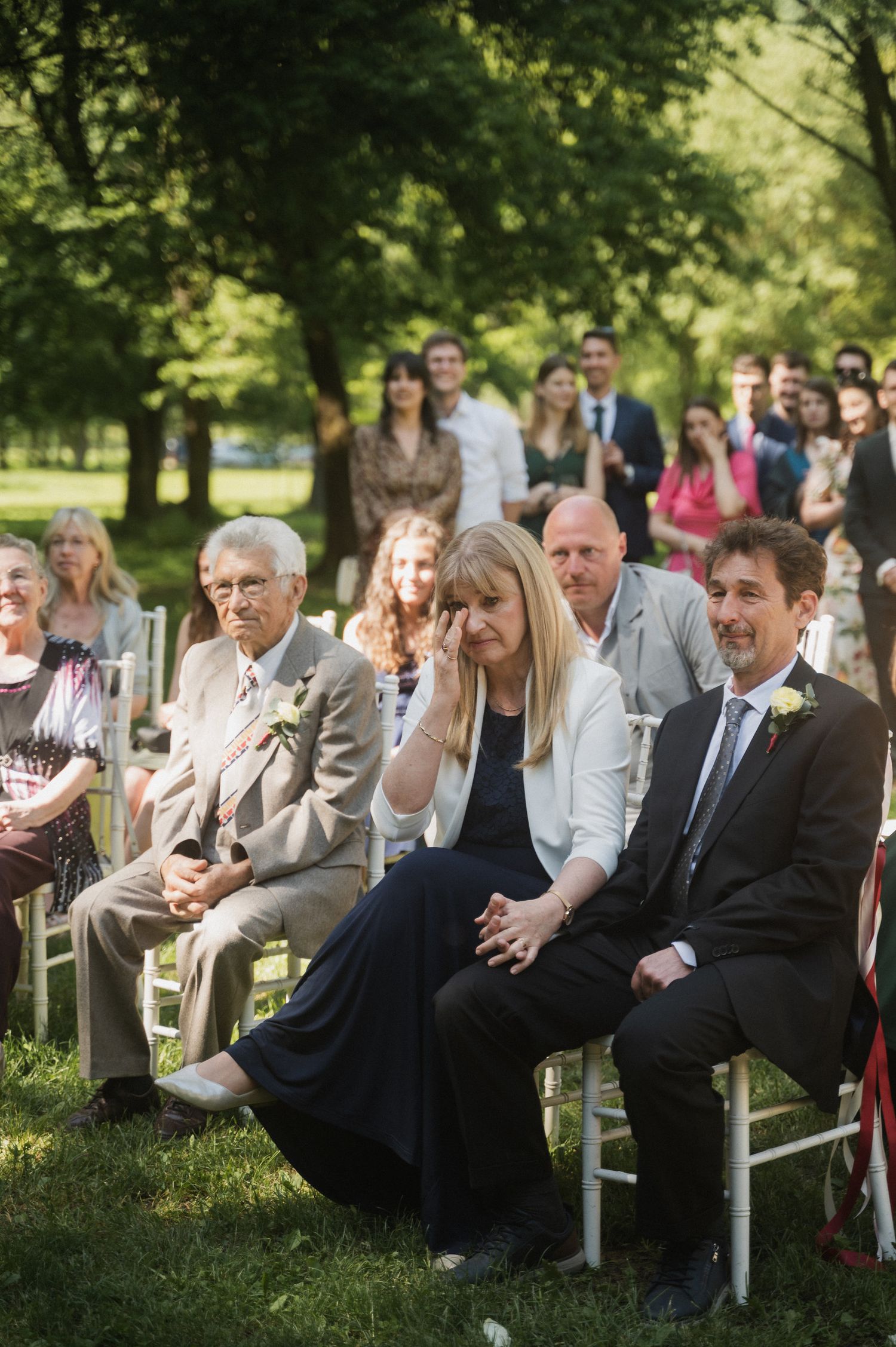 Wedding guests seated in white chairs outdoors react with laughter during an outdoor ceremony in a green park setting.