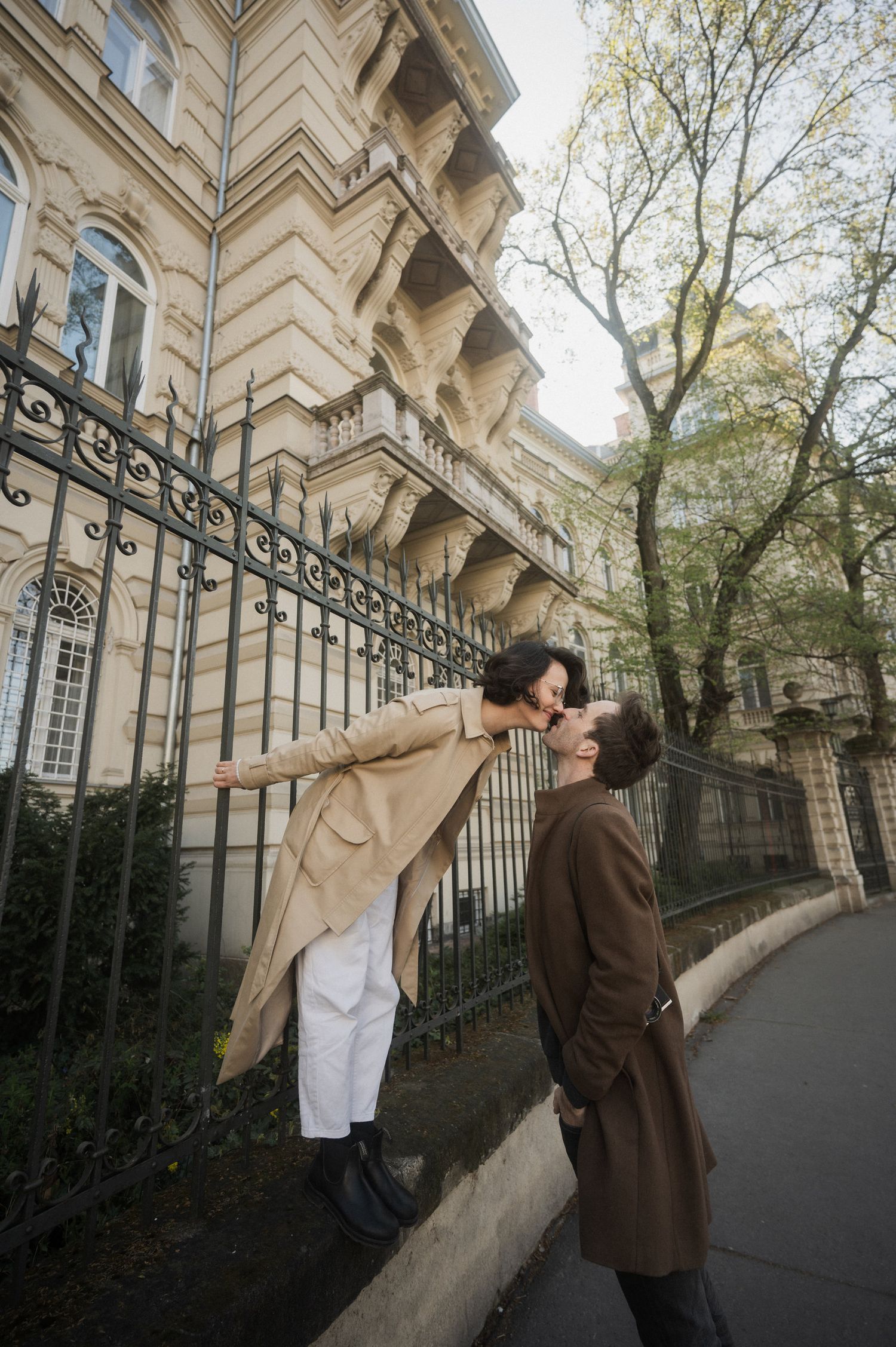 Two people walk along an ornate European building exterior with wrought iron fencing and classical architecture details.