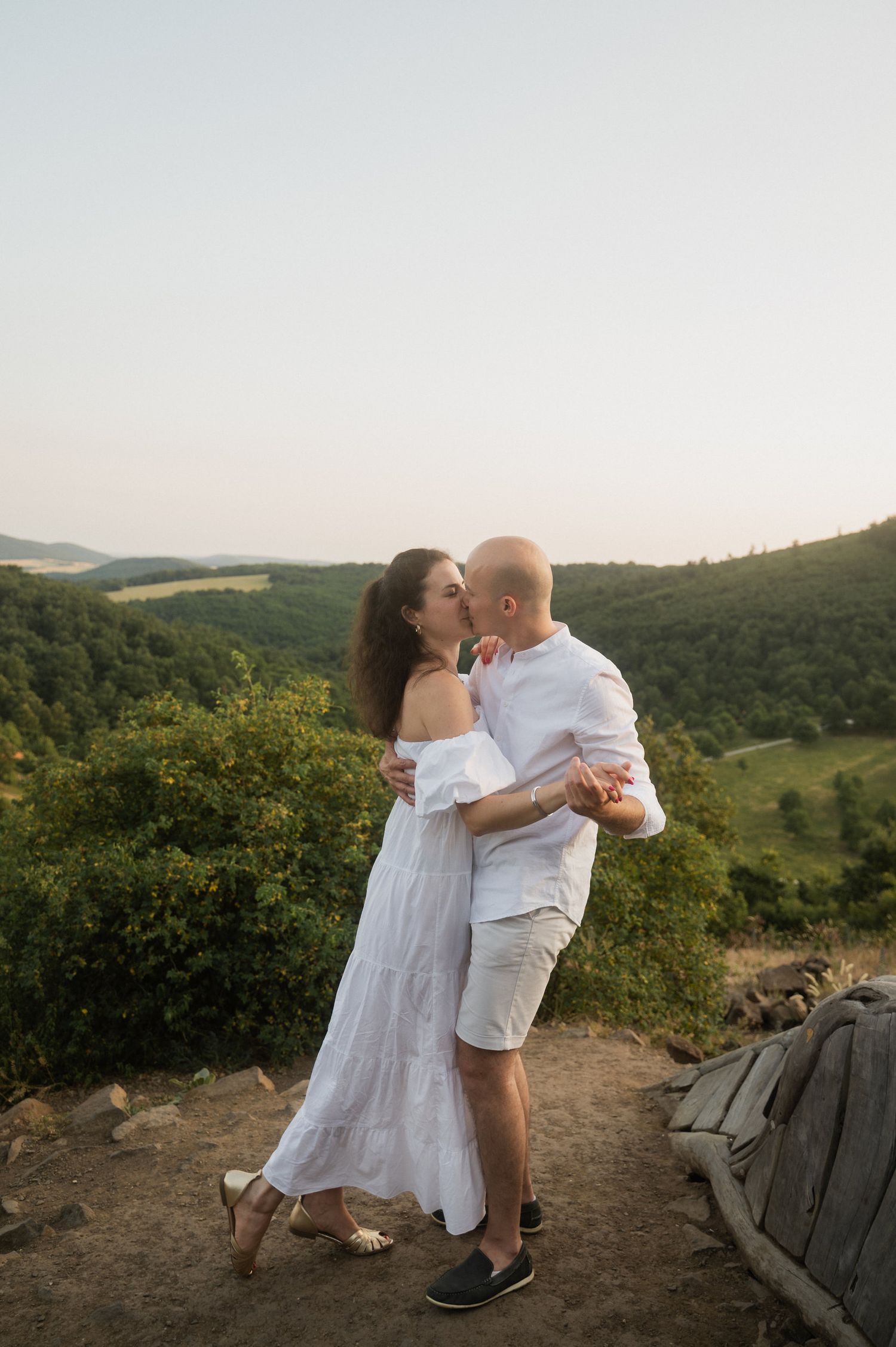 A couple in white outfits dancing together on a hilltop at sunset with mountains in the background.