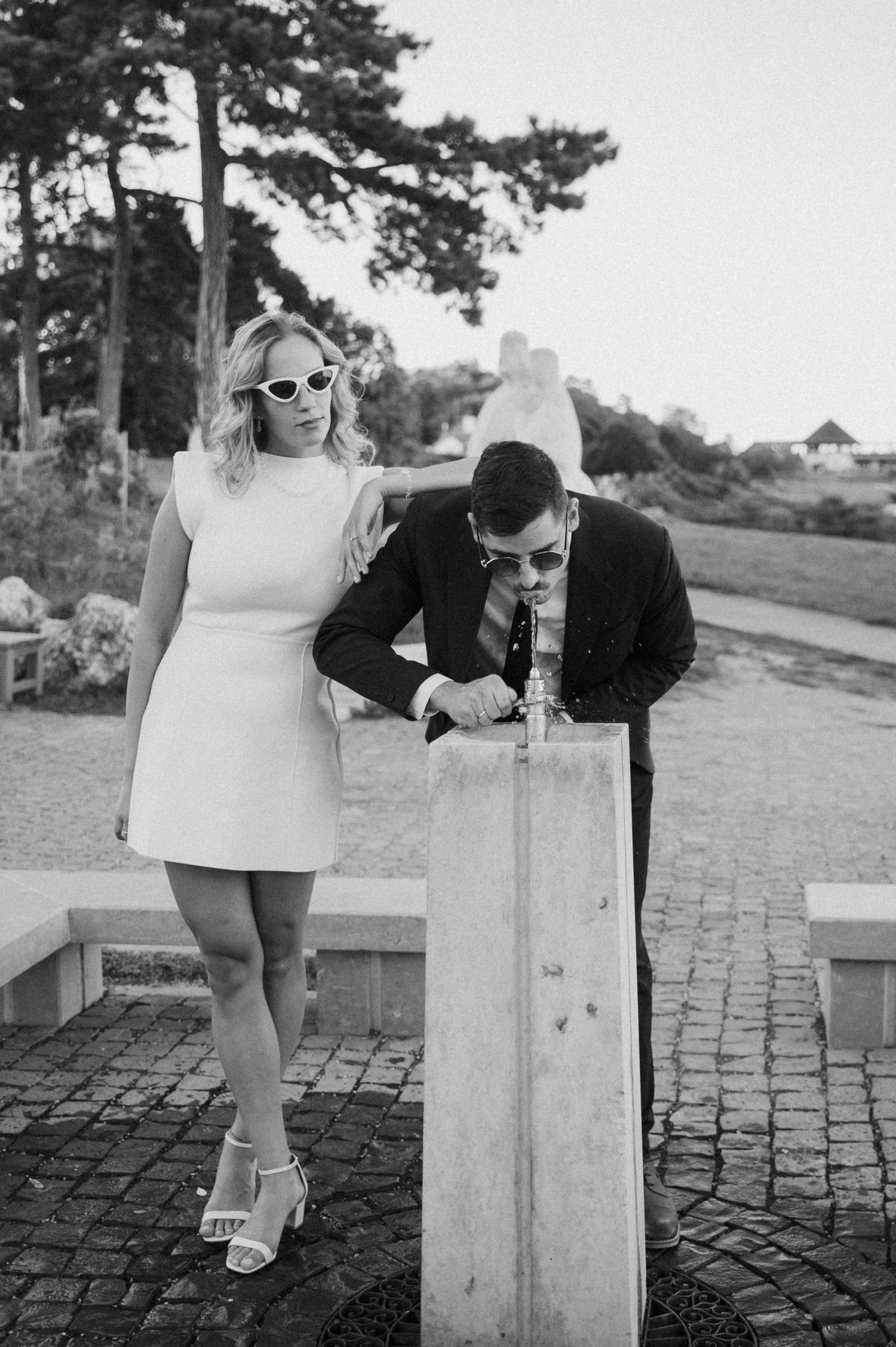 A stylish couple in sunglasses pose by a stone pillar, with the woman in a white mini dress and man in dark suit.
