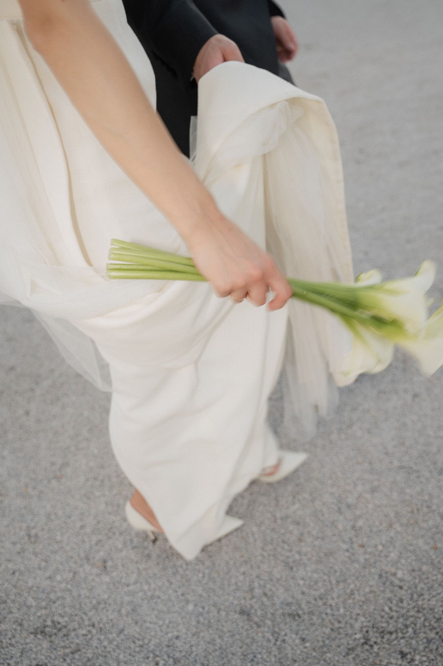 Detail shot of white wedding dress and calla lily bouquet against stone ground.