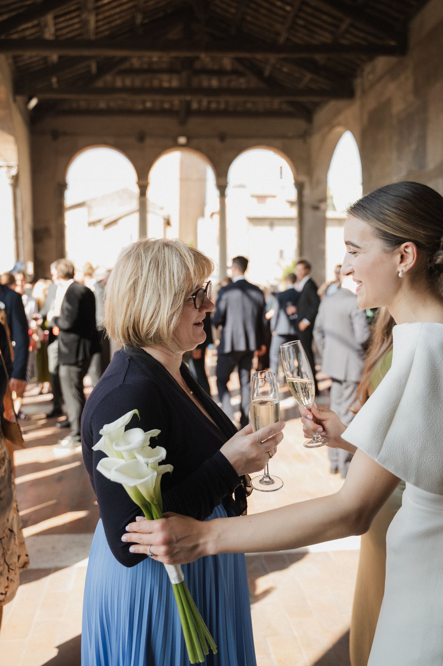 Wedding party members exchange flowers and greetings under a covered stone walkway.
